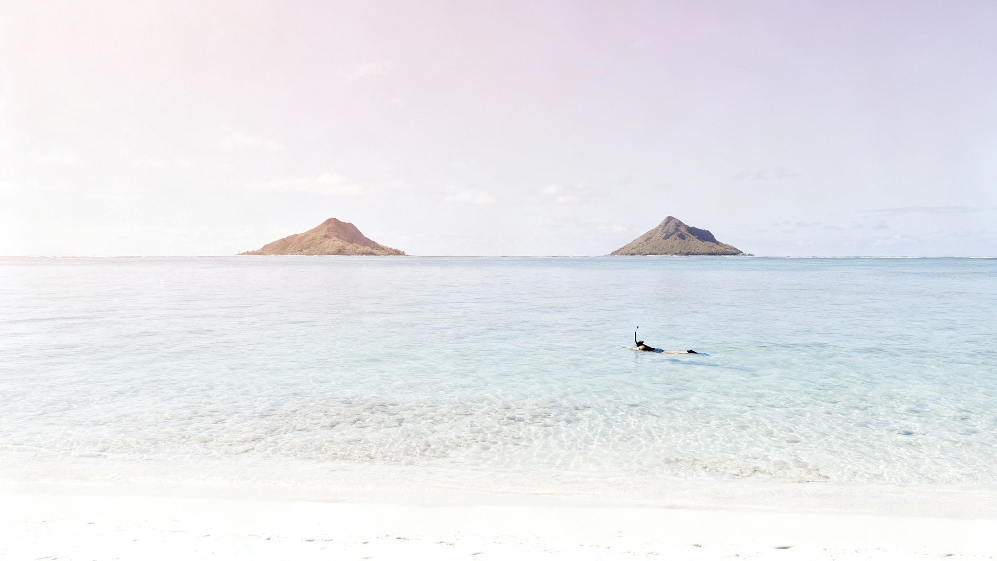 A person snorkeling in crystal clear ocean water with two volcanic islands in the background.