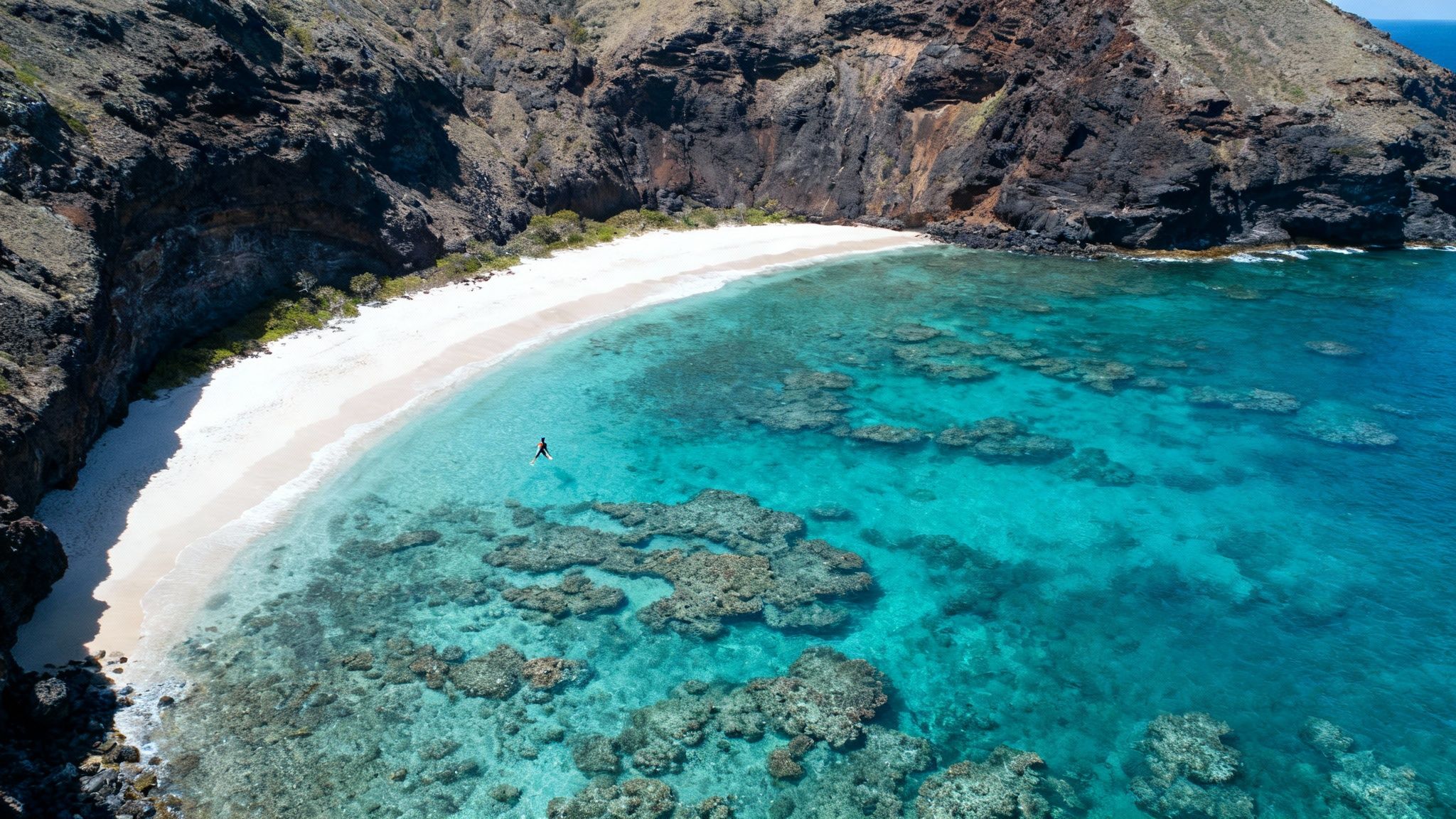 Stunning aerial view of a tropical beach with clear turquoise water, coral reefs, and a paddleboarder.