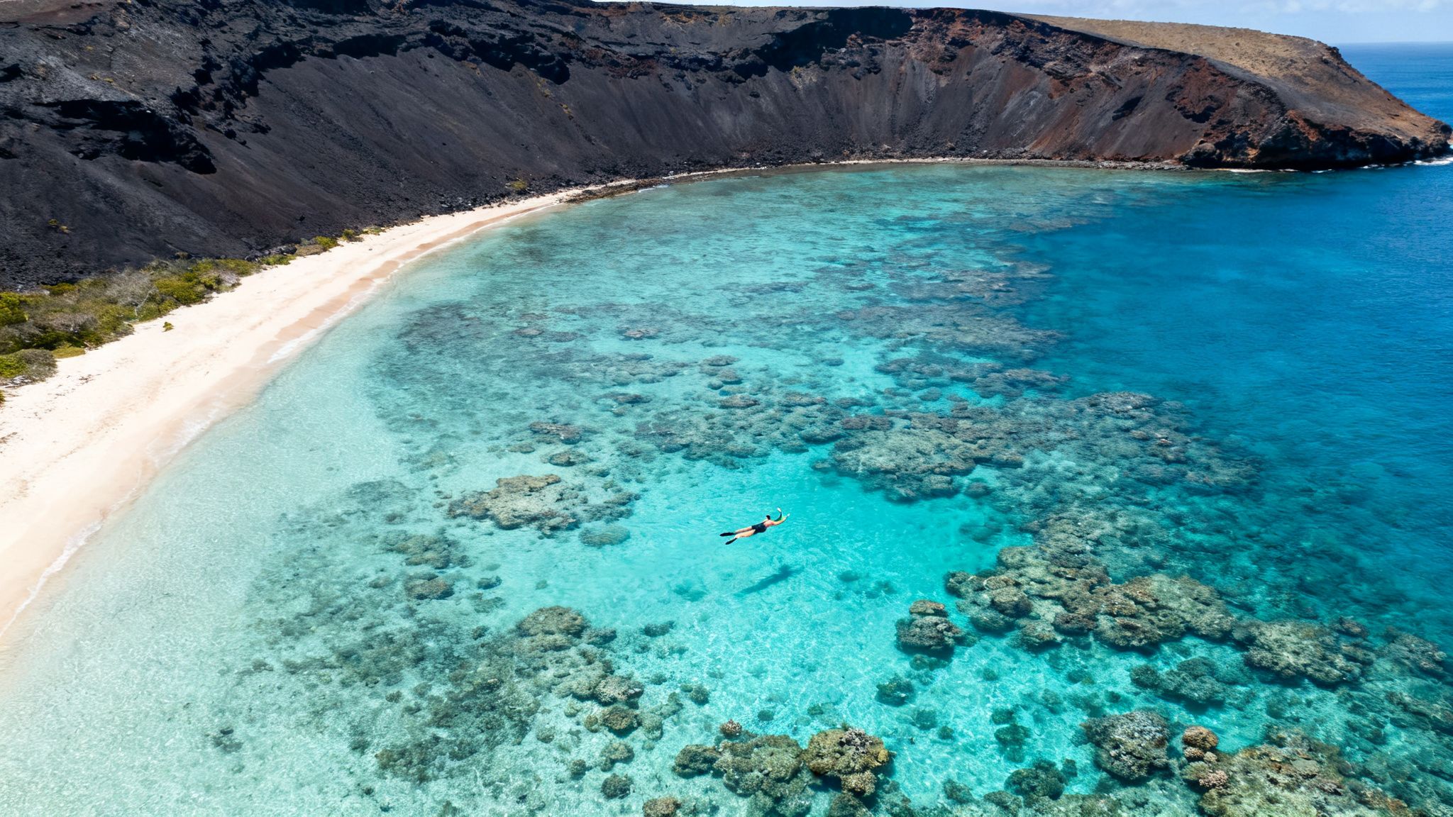 Aerial view of a person snorkeling over coral reefs in a turquoise bay with a black volcanic crater.