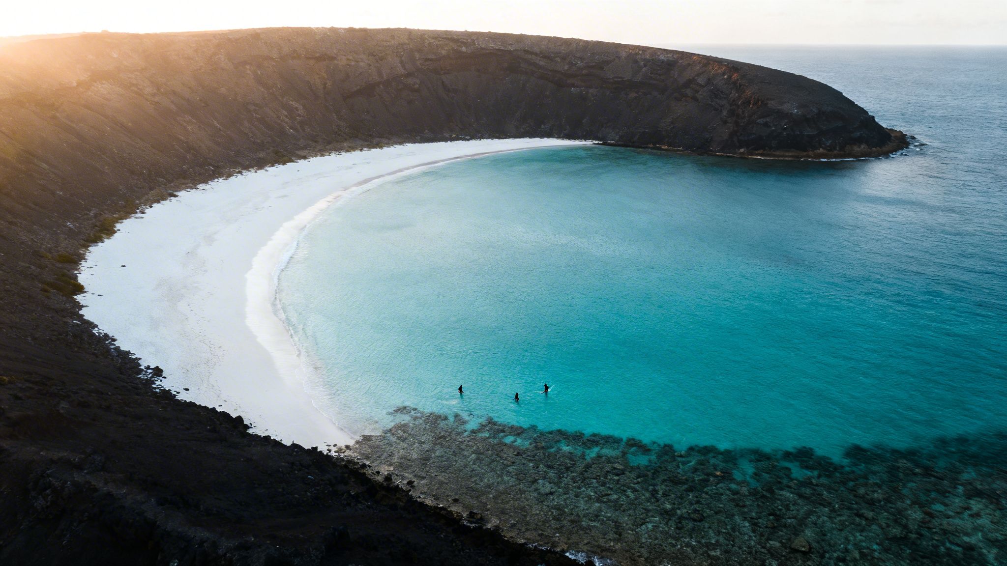 Aerial view of a secluded white sand beach with turquoise water and volcanic cliffs at sunset.