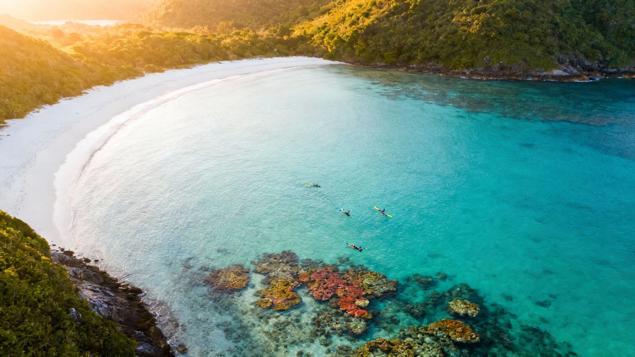 Stunning aerial view of a tropical bay with white sandy beach, turquoise water, green hills, and kayakers.