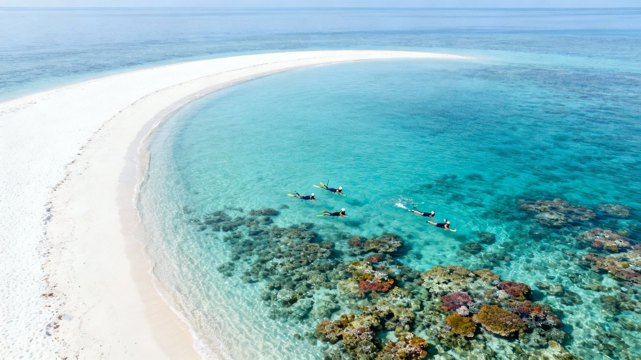 Aerial view of a pristine white sandbar, clear turquoise ocean, and snorkelers exploring coral reefs.