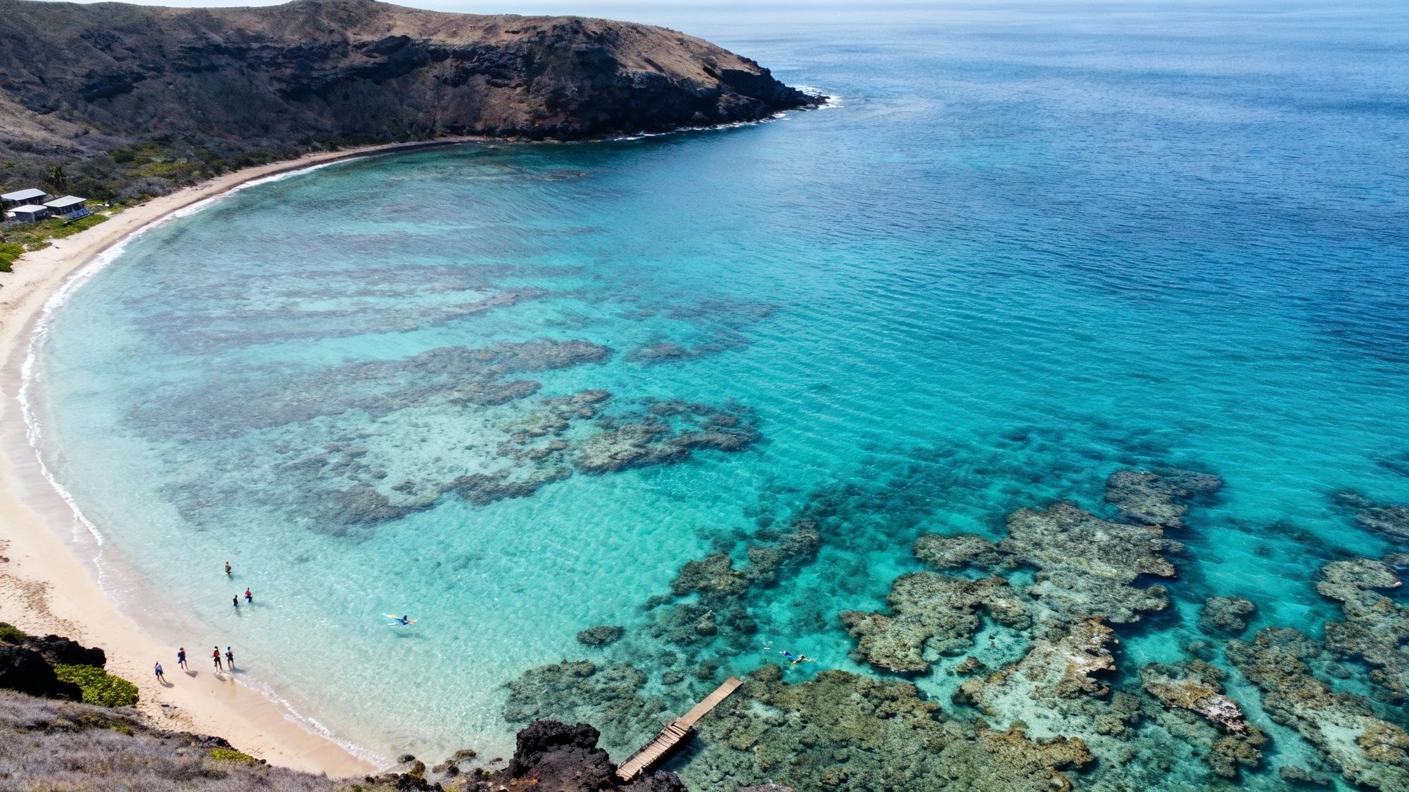 Aerial view of a beautiful turquoise bay with a sandy beach, coral reefs, and people snorkeling.