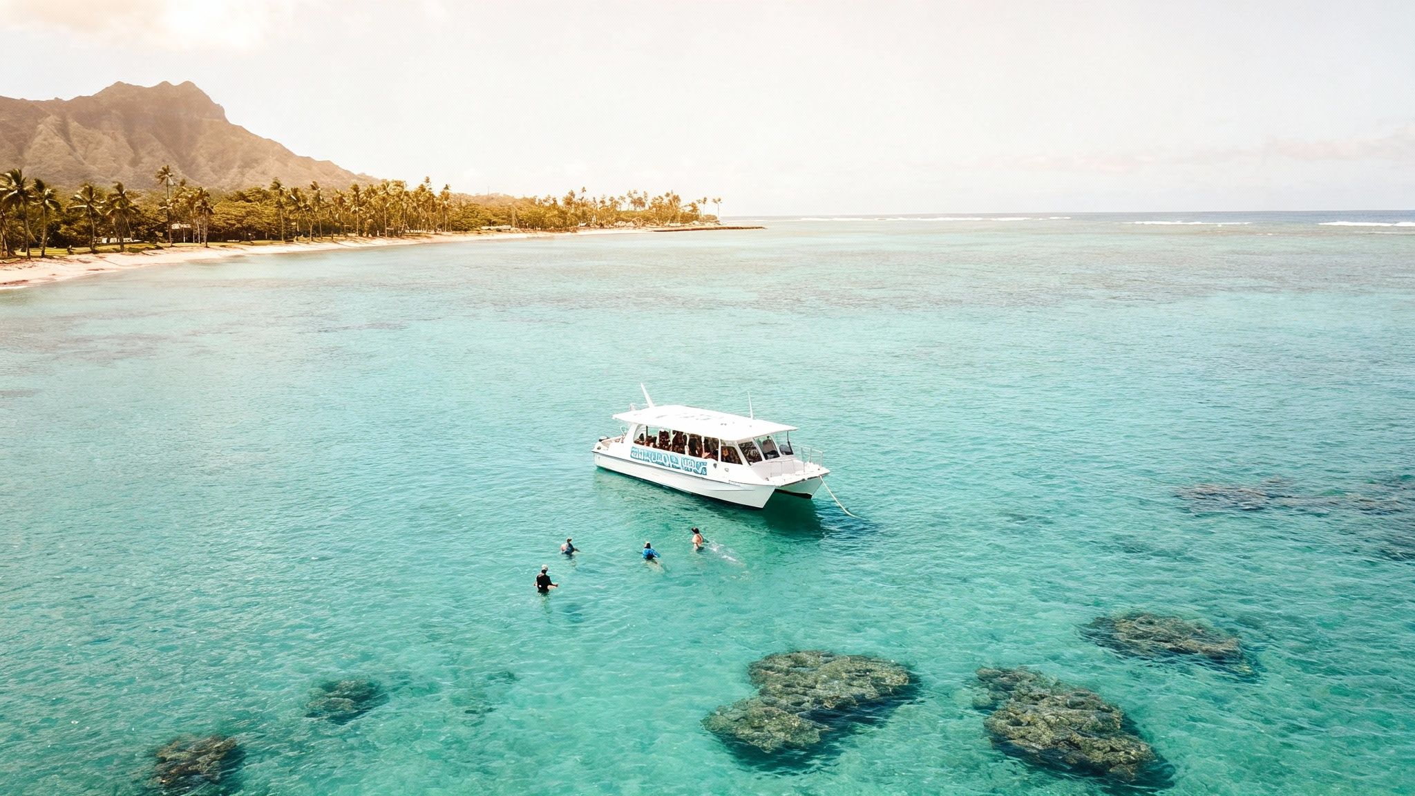 An aerial view of a white boat with snorkelers in clear turquoise water near a tropical beach and mountain.