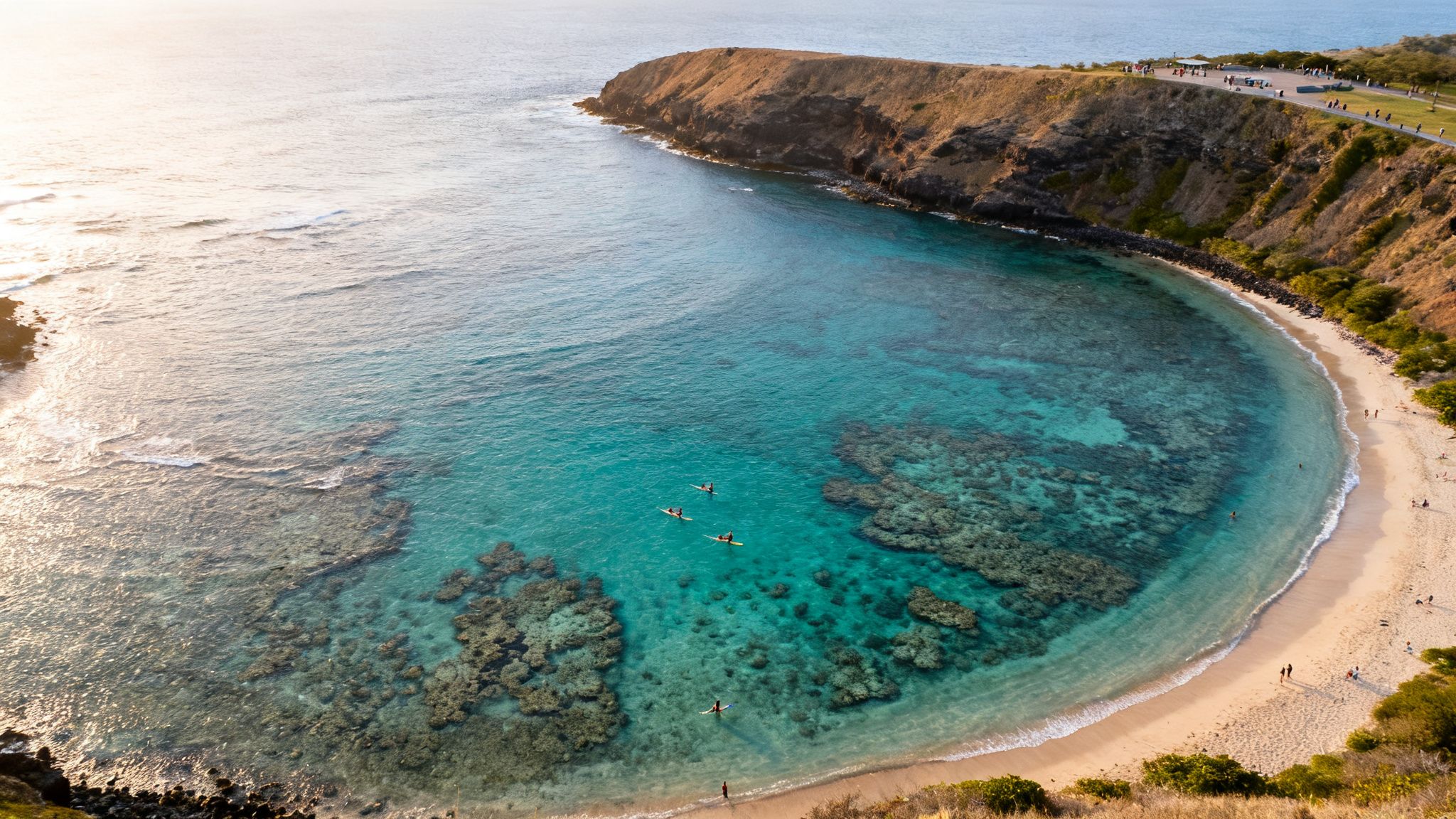 Aerial view of a clear turquoise bay with visible coral reefs, paddleboarders, a sandy beach, and cliffside path.