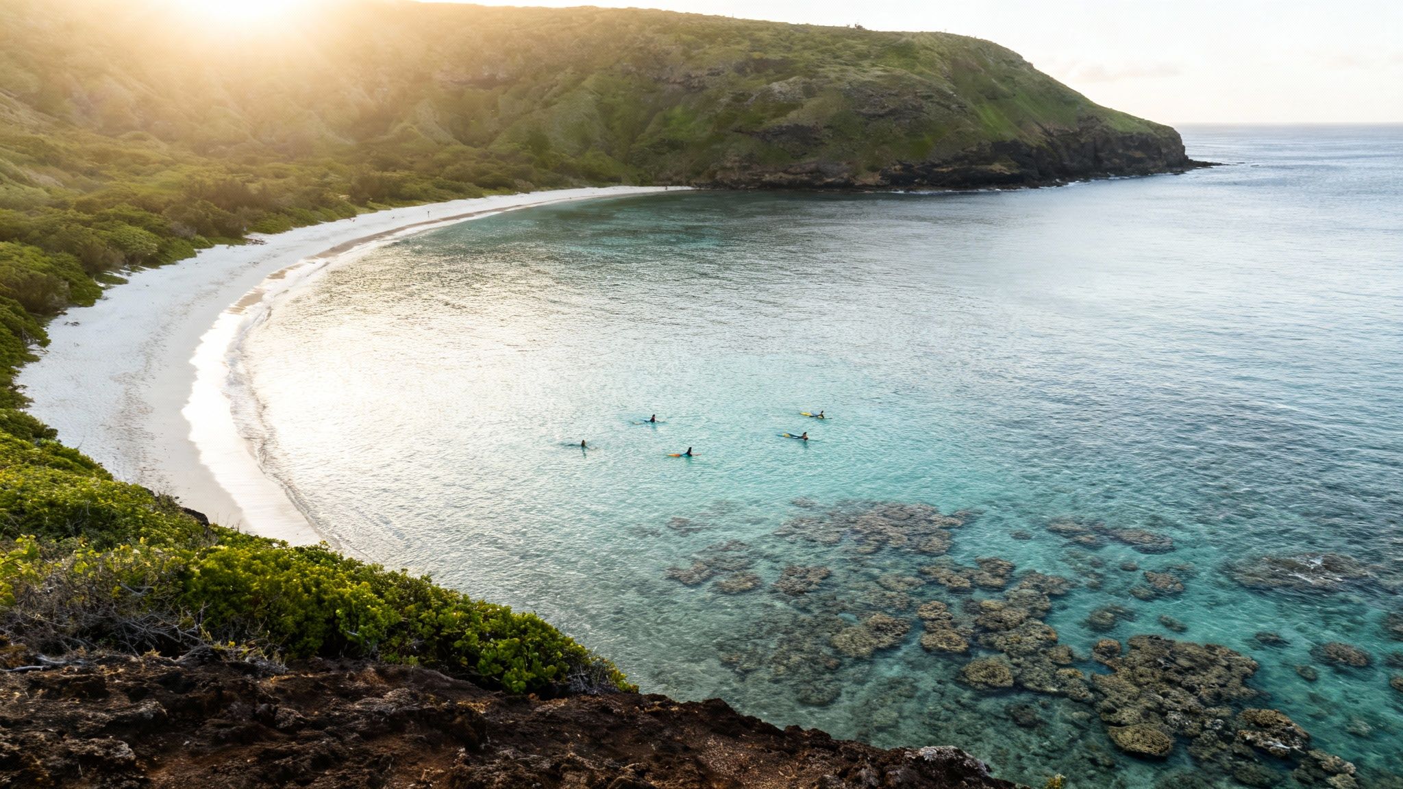 Aerial view of a sunny Hawaiian bay with a white sand beach, green hills, and people on surfboards.