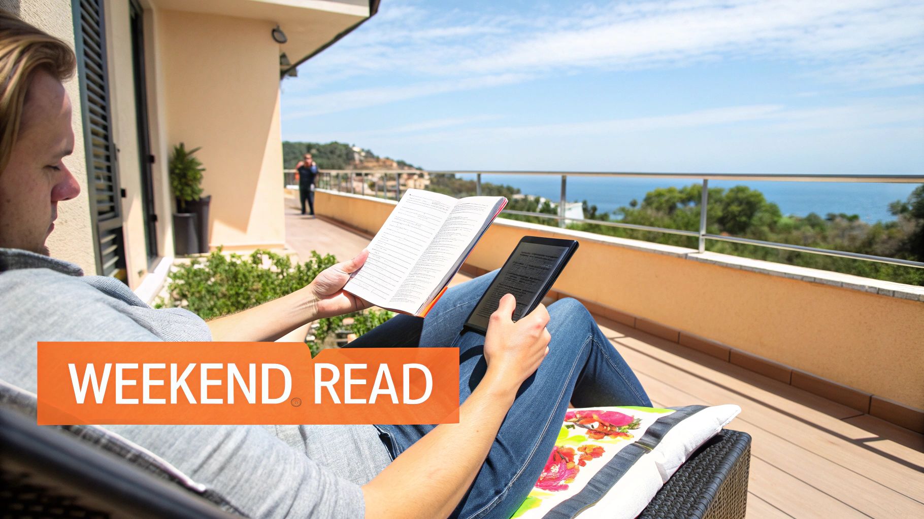A person reads a physical book and an e-reader on a sunny balcony overlooking the ocean.