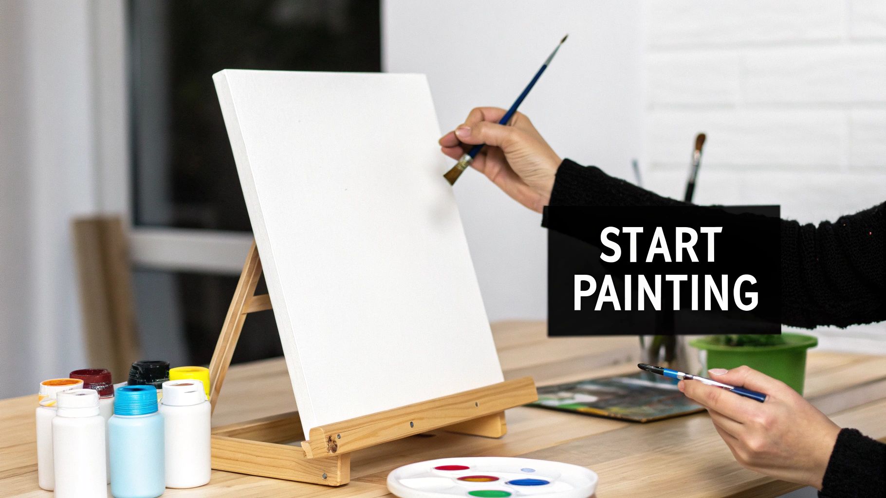 Person's hands holding paintbrushes, about to paint on a blank canvas with colorful acrylic paints.