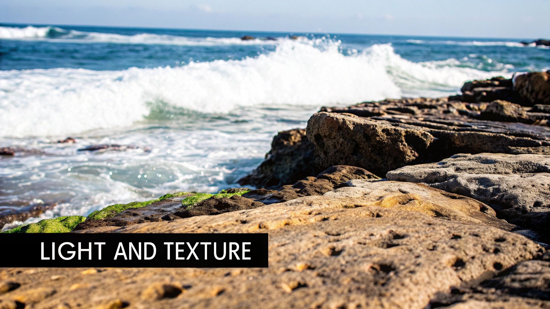 Textured coastal rocks with green moss, ocean waves crashing, and sunlight highlighting details.