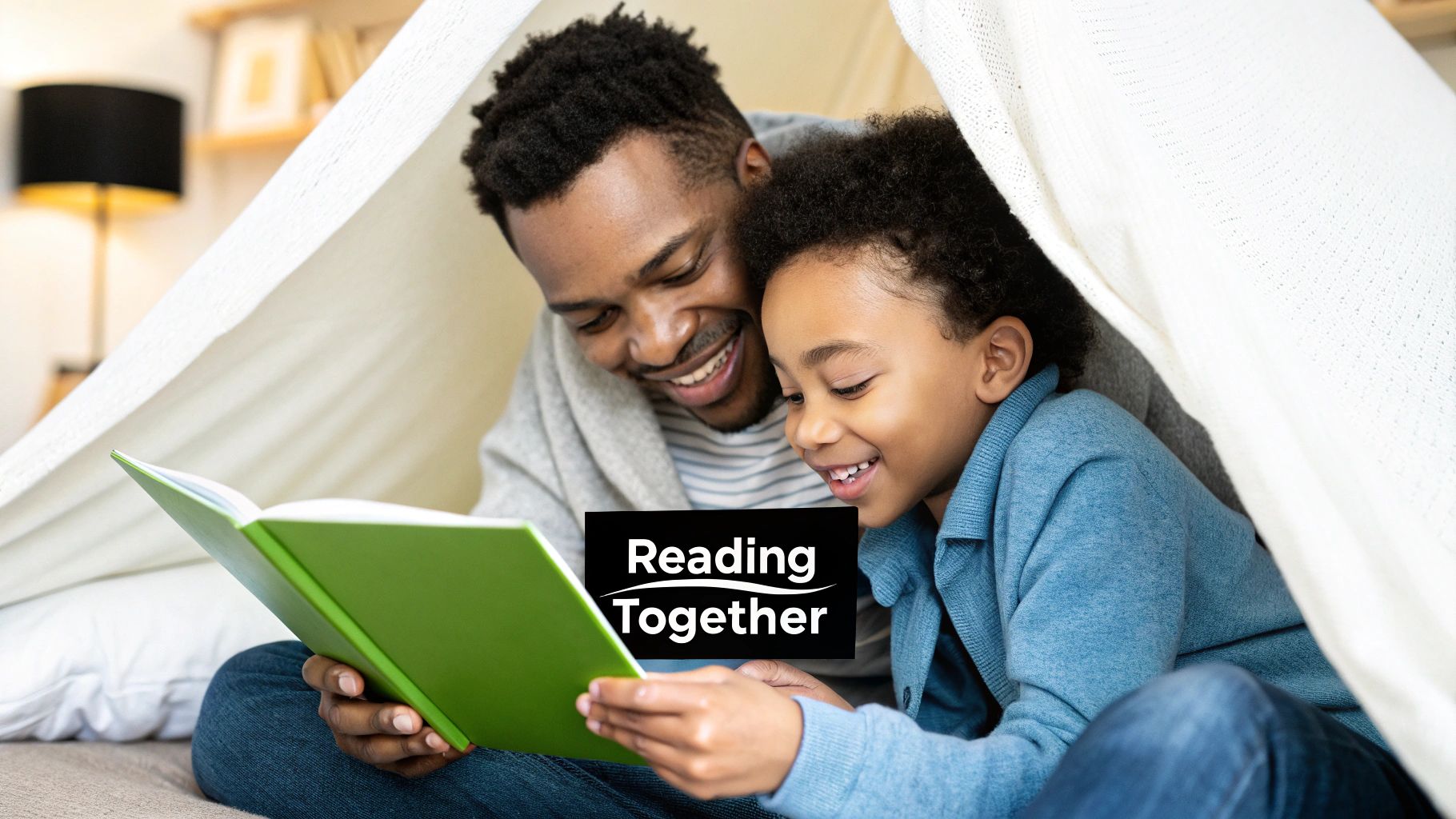 A smiling man and child reading a green book together in a cozy blanket fort.