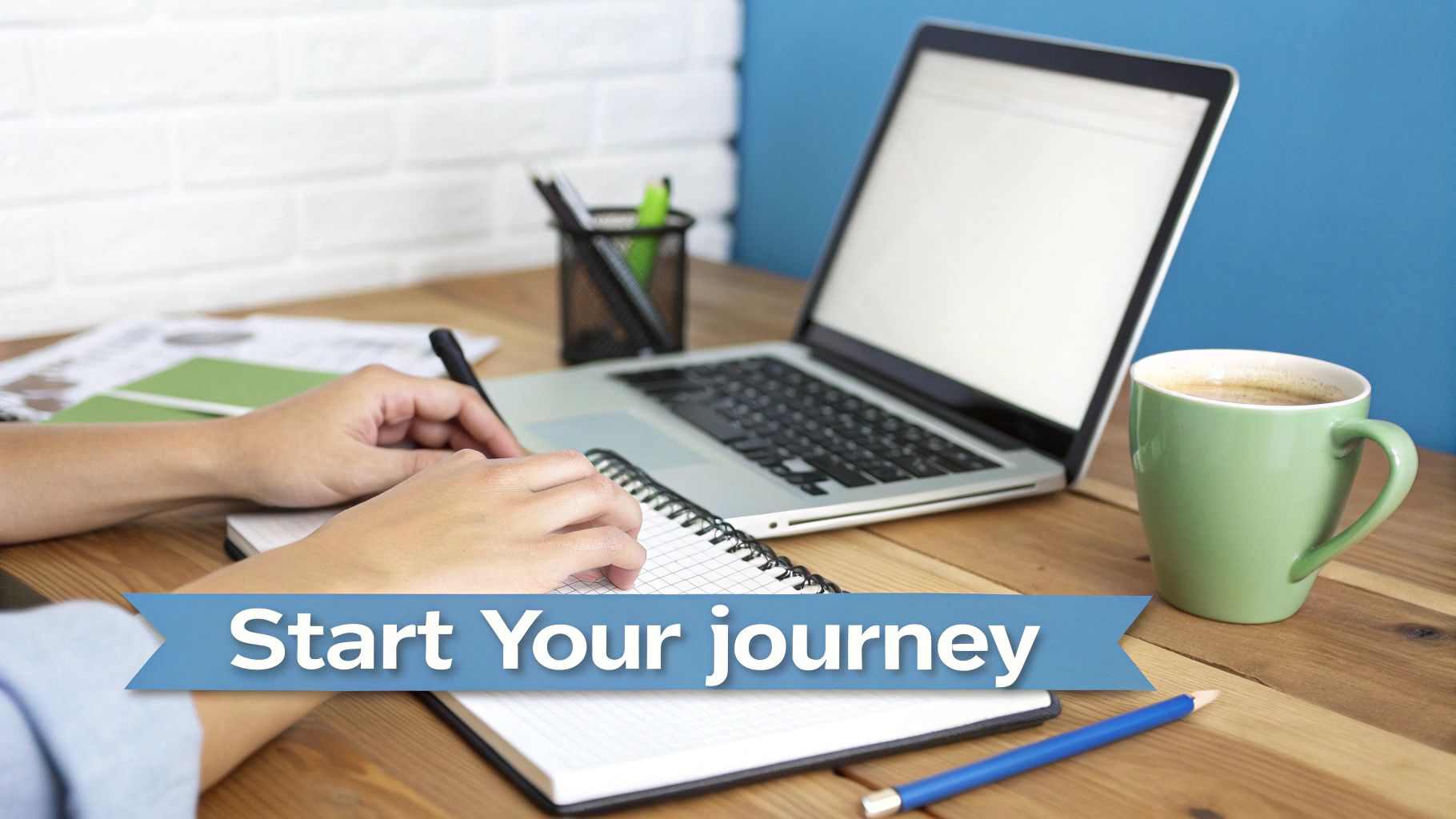 A person's hands writing in a notebook on a wooden desk with a laptop, coffee, and a 'Start Your journey' banner.