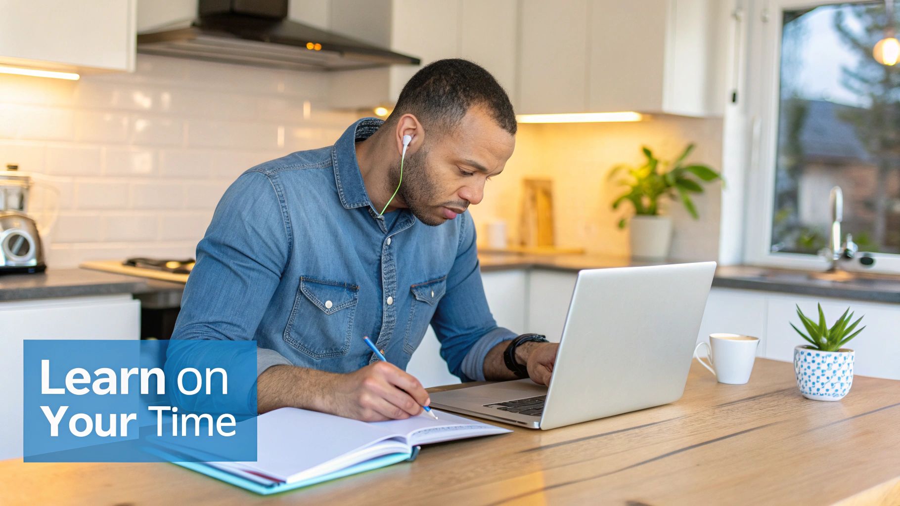 A man in a denim shirt uses a laptop and writes in a notebook at his kitchen counter, studying or working remotely.