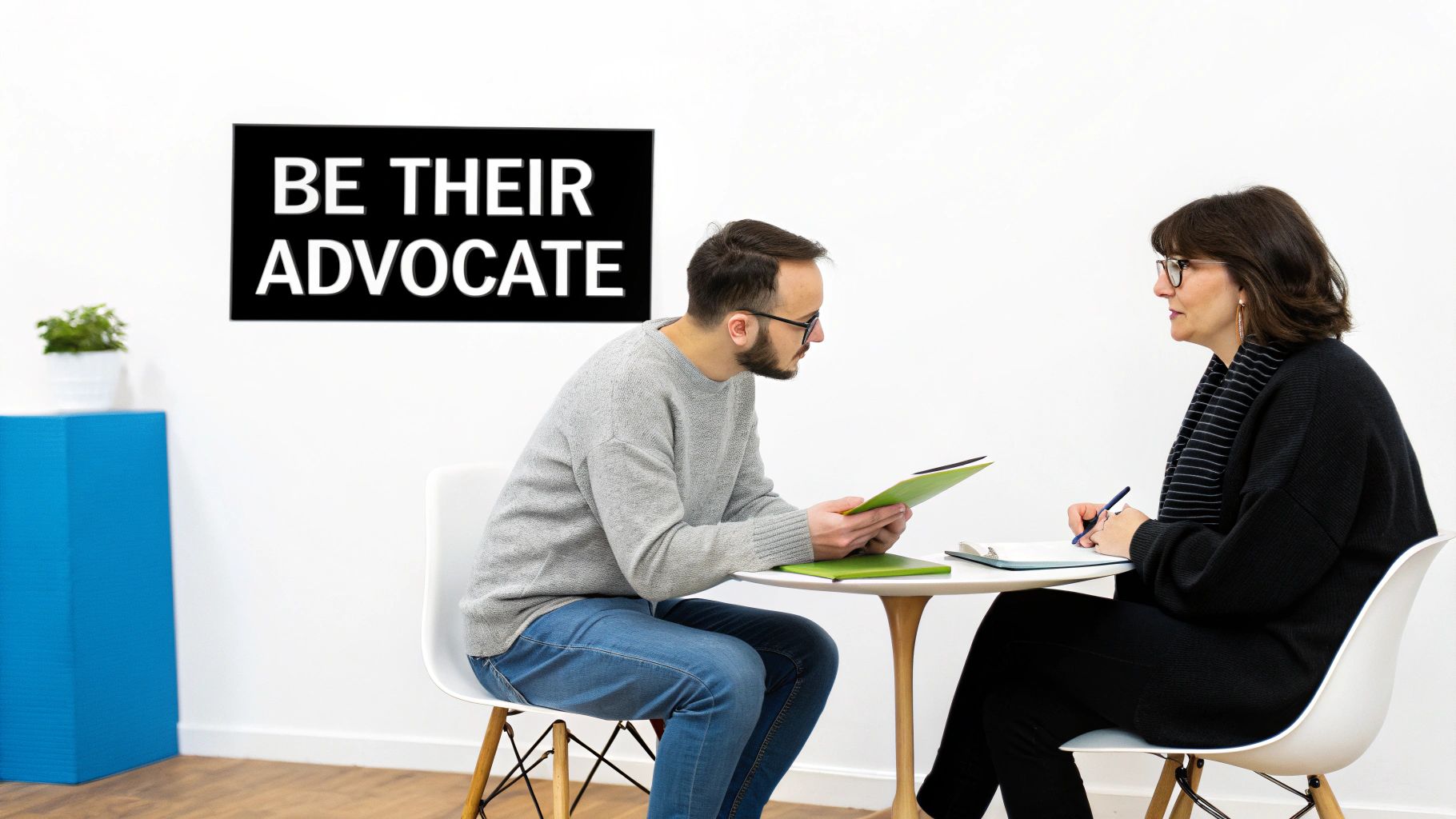 Two people, a man and a woman, discussing or consulting at a table, with 'BE THEIR ADVOCATE' on the wall.