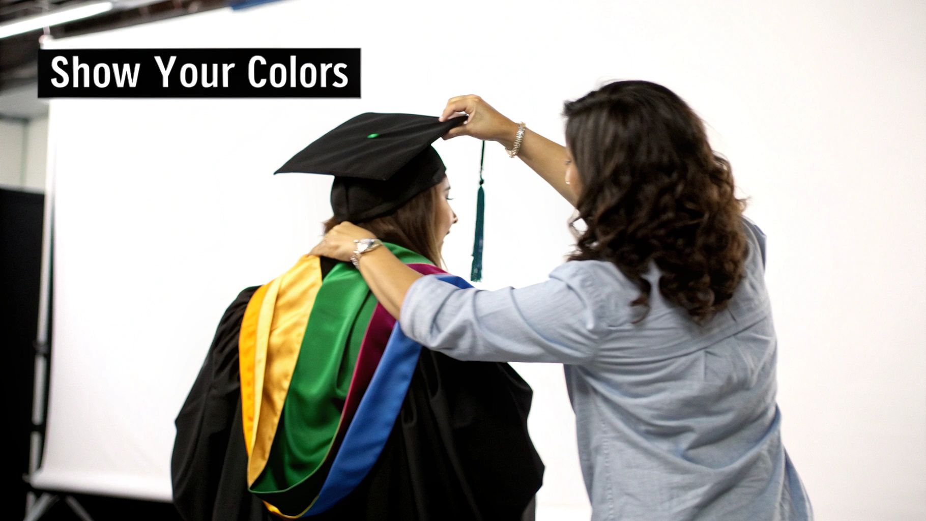 A person in a graduation cap, gown, and colorful academic hood being helped by another.