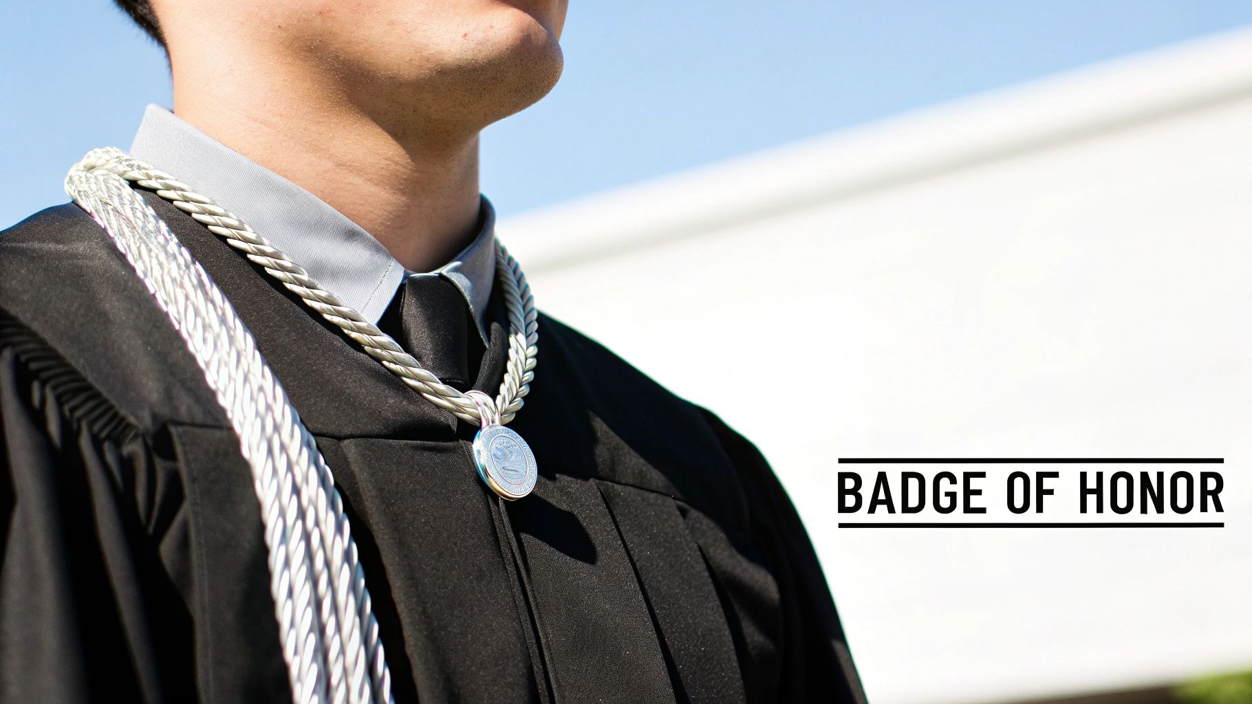 A student in black graduation robes and a grey shirt wears silver honor cords and a medallion.