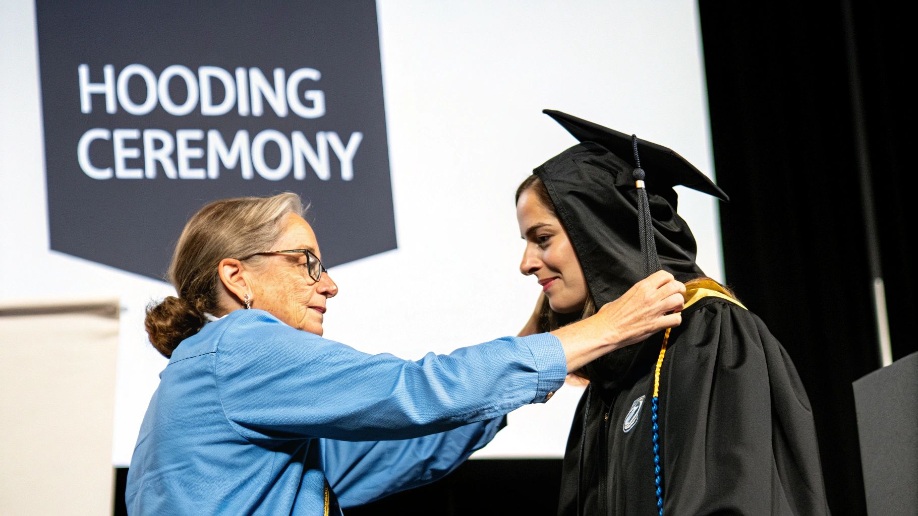 An older woman helps a smiling graduate wear her academic hood at a hooding ceremony.