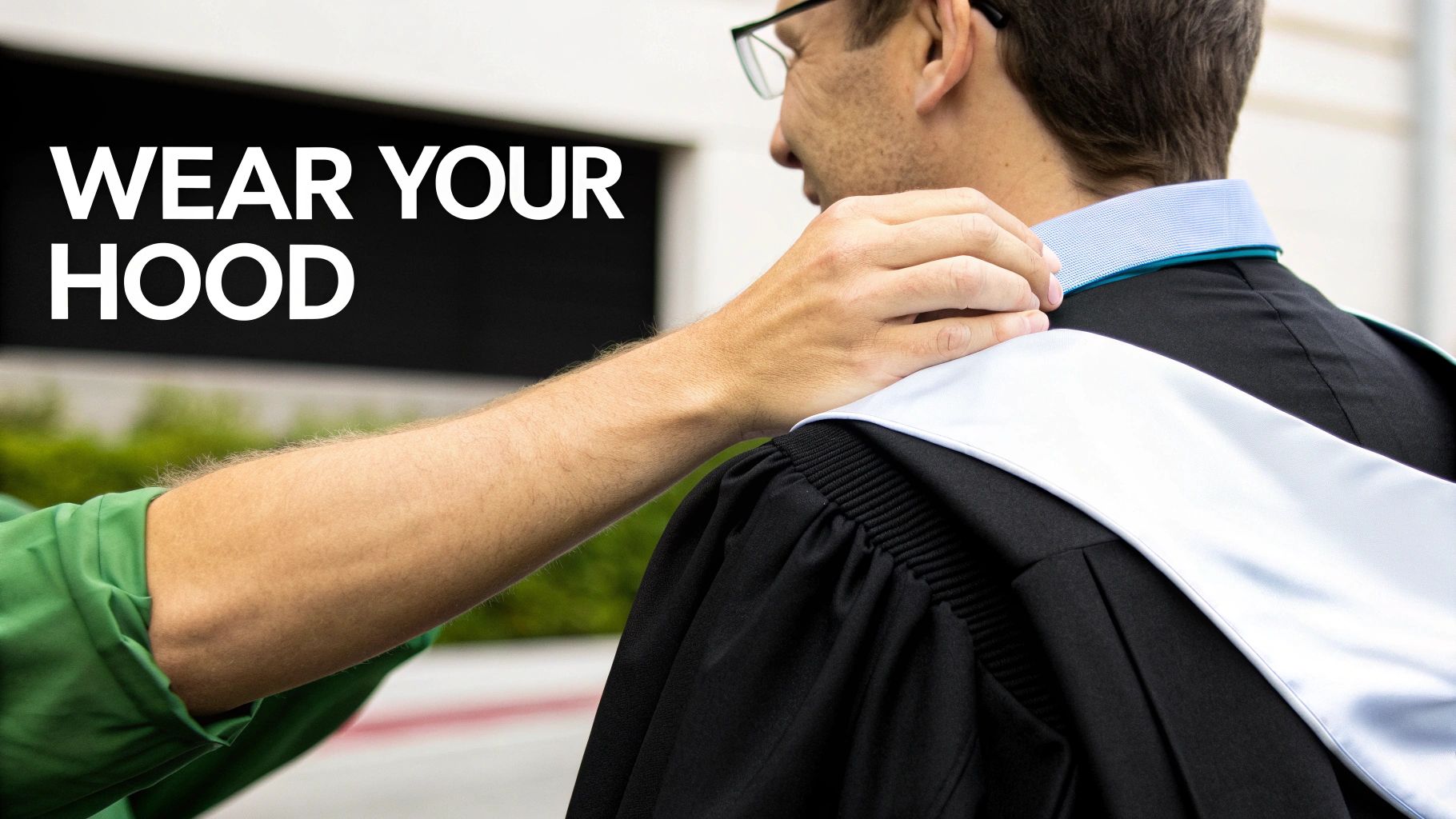 A person in a black graduation gown and light blue hood having it adjusted by another person, with text 'WEAR YOUR HOOD'.