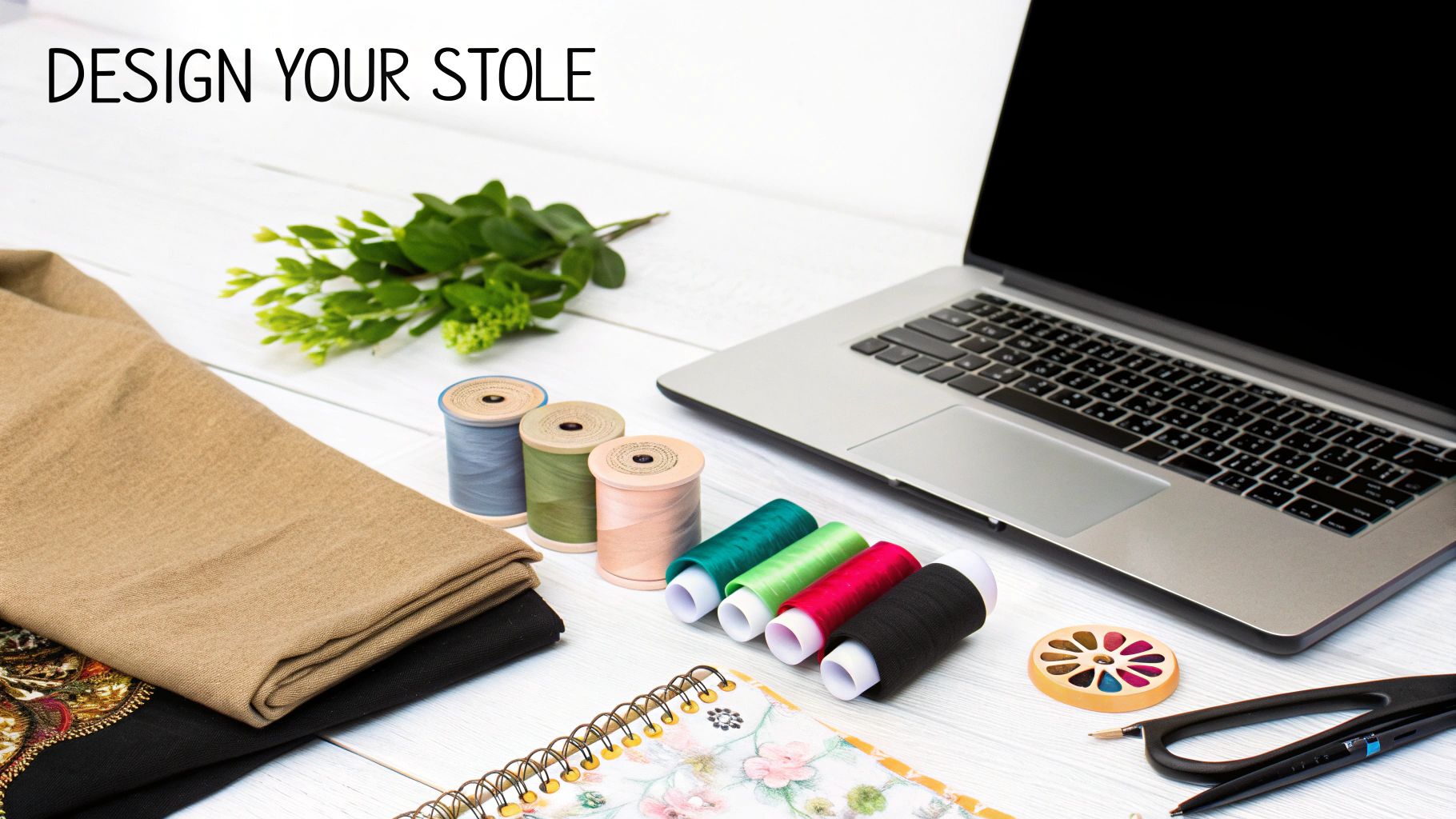 Flat lay of a laptop, colorful threads, fabrics, and design tools on a white wooden desk.