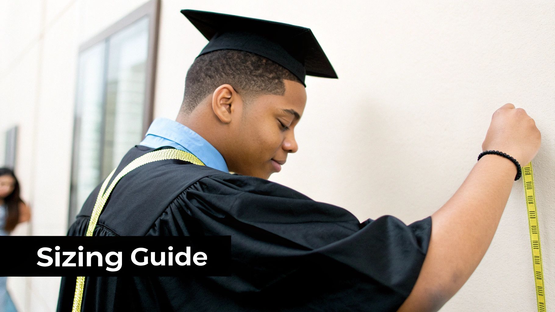 A young graduate in cap and gown measures their height against a wall with a yellow tape, with 'Sizing Guide' text.