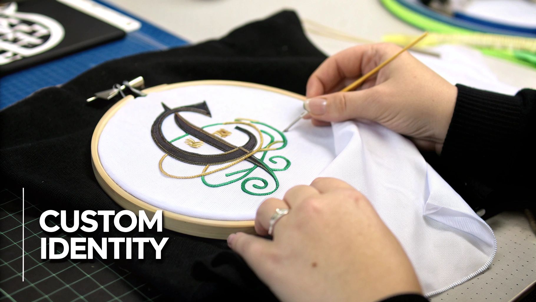 Close-up of hands embroidering a custom monogram 'C' with green and gold thread on white fabric.
