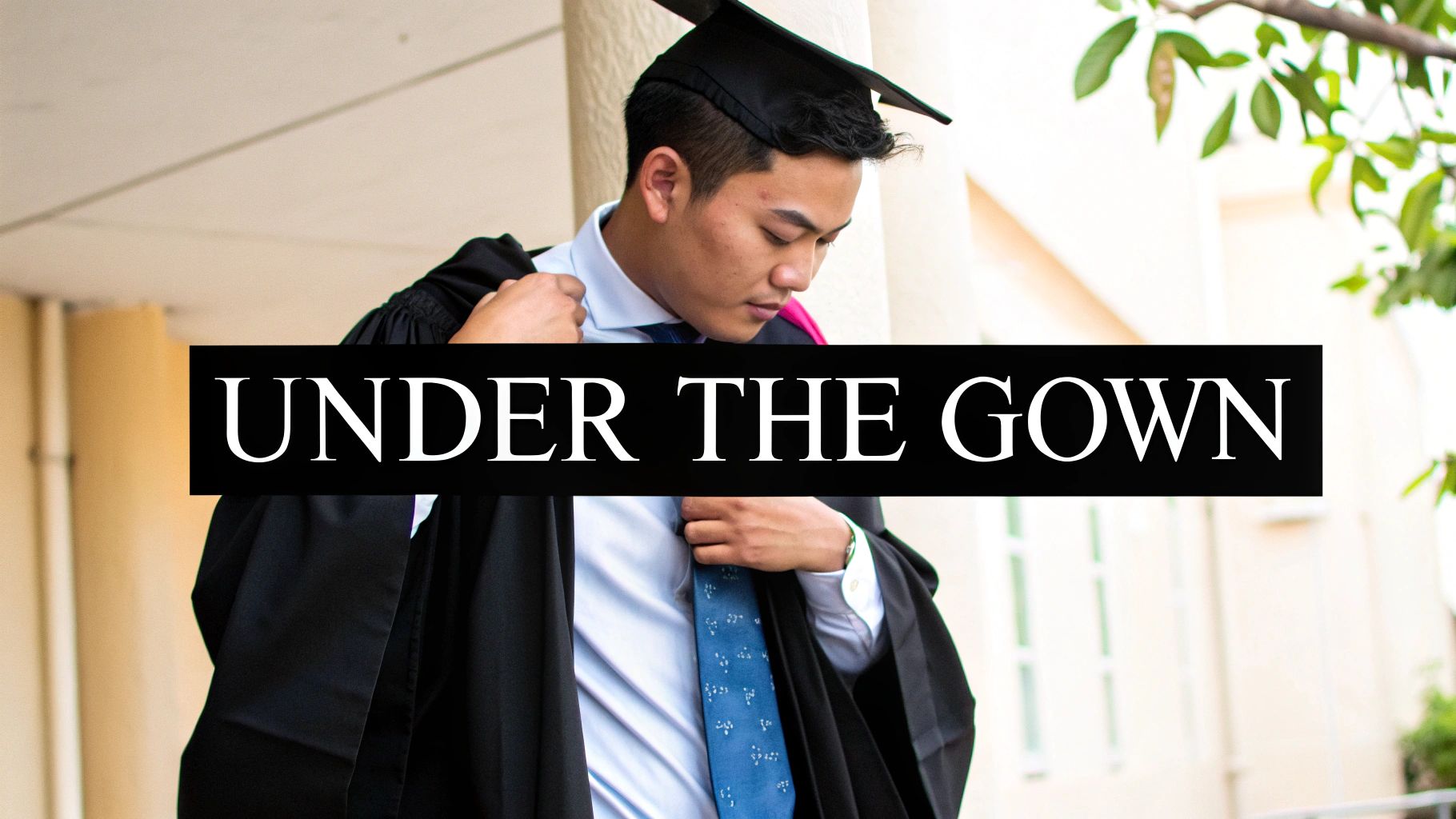 A male graduate in a cap and gown adjusting his blue shirt and patterned tie.