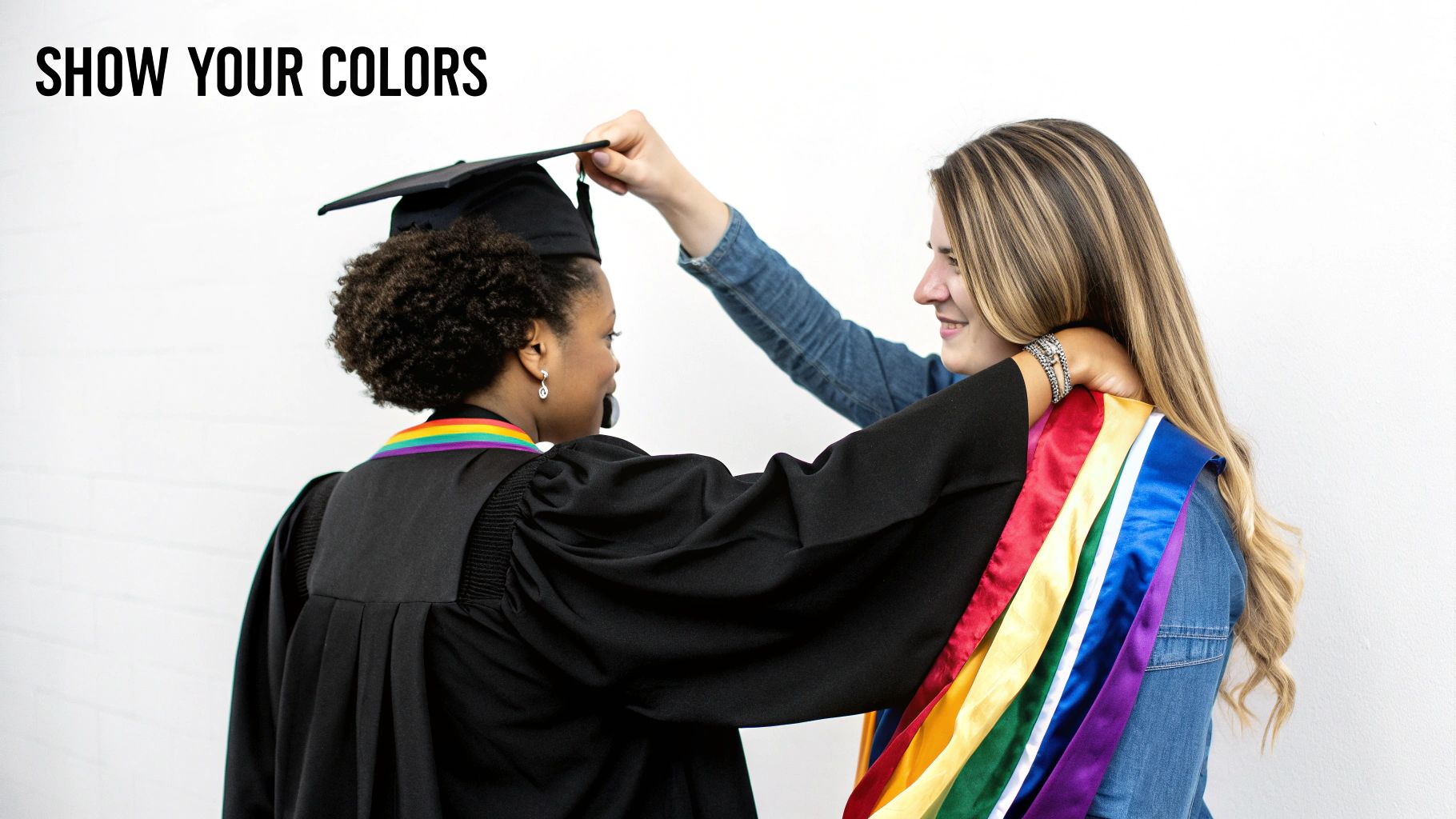 Two smiling women, one in a graduation cap and gown with a rainbow stole, the other helping her, symbolizing LGBTQ+ pride.