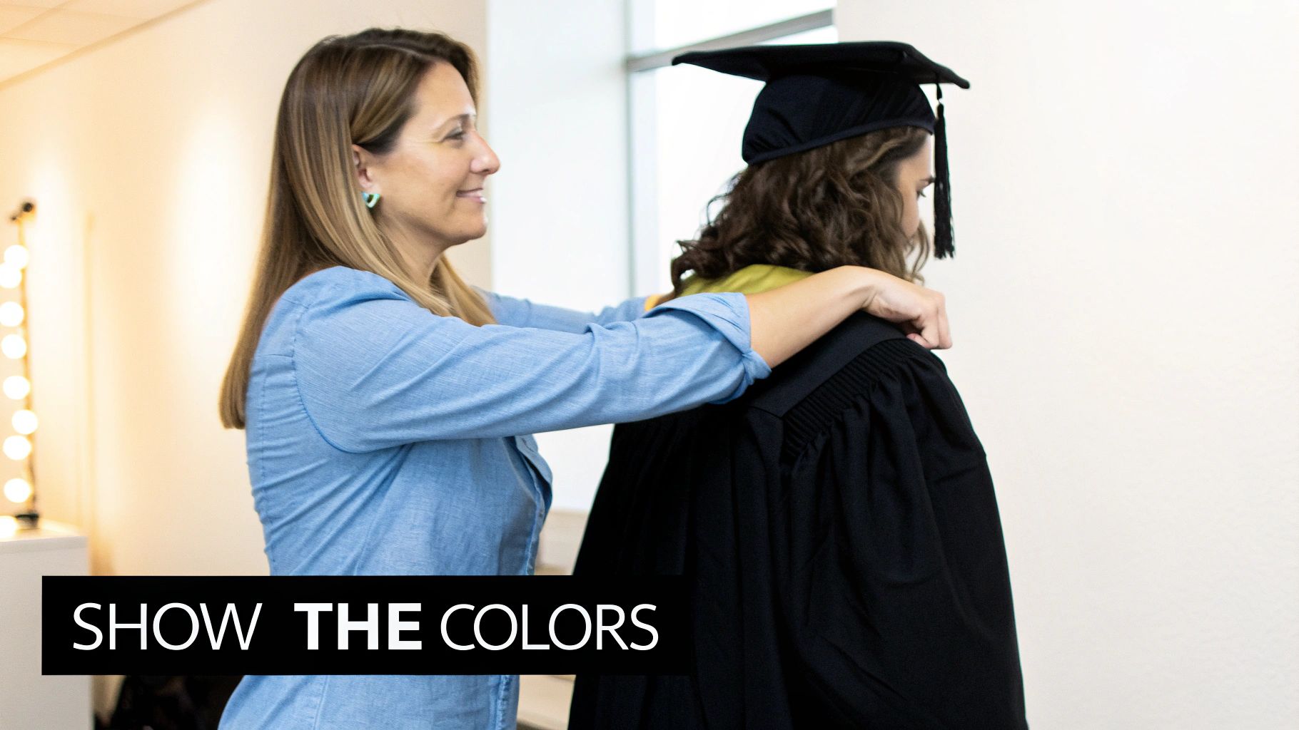A woman in a blue shirt helps another person put on a black graduation gown and academic hood.