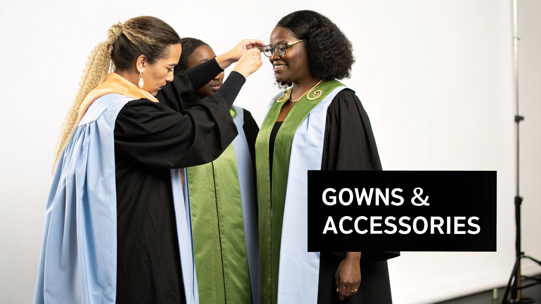 Three women in academic gowns and colorful stoles, with one adjusting another's glasses, ready for graduation.