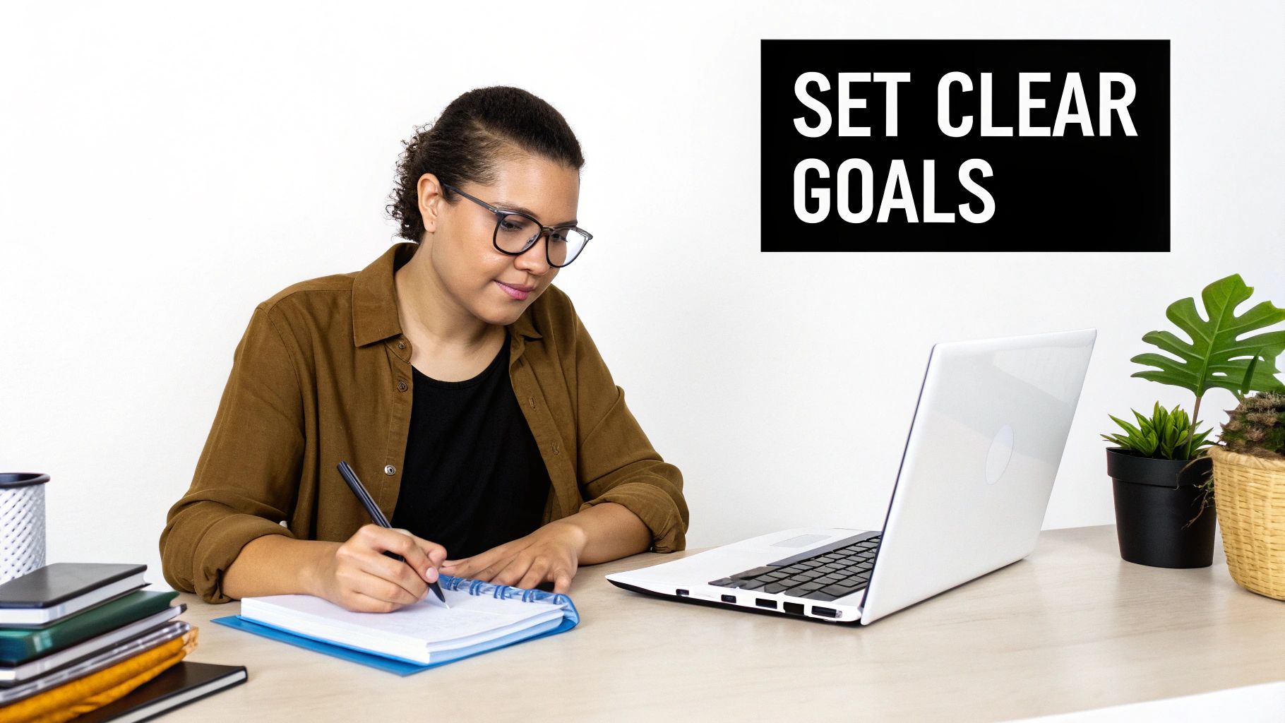 A person with glasses writing in a notebook at a desk with a laptop, books, and plants. Text: SET CLEAR GOALS.