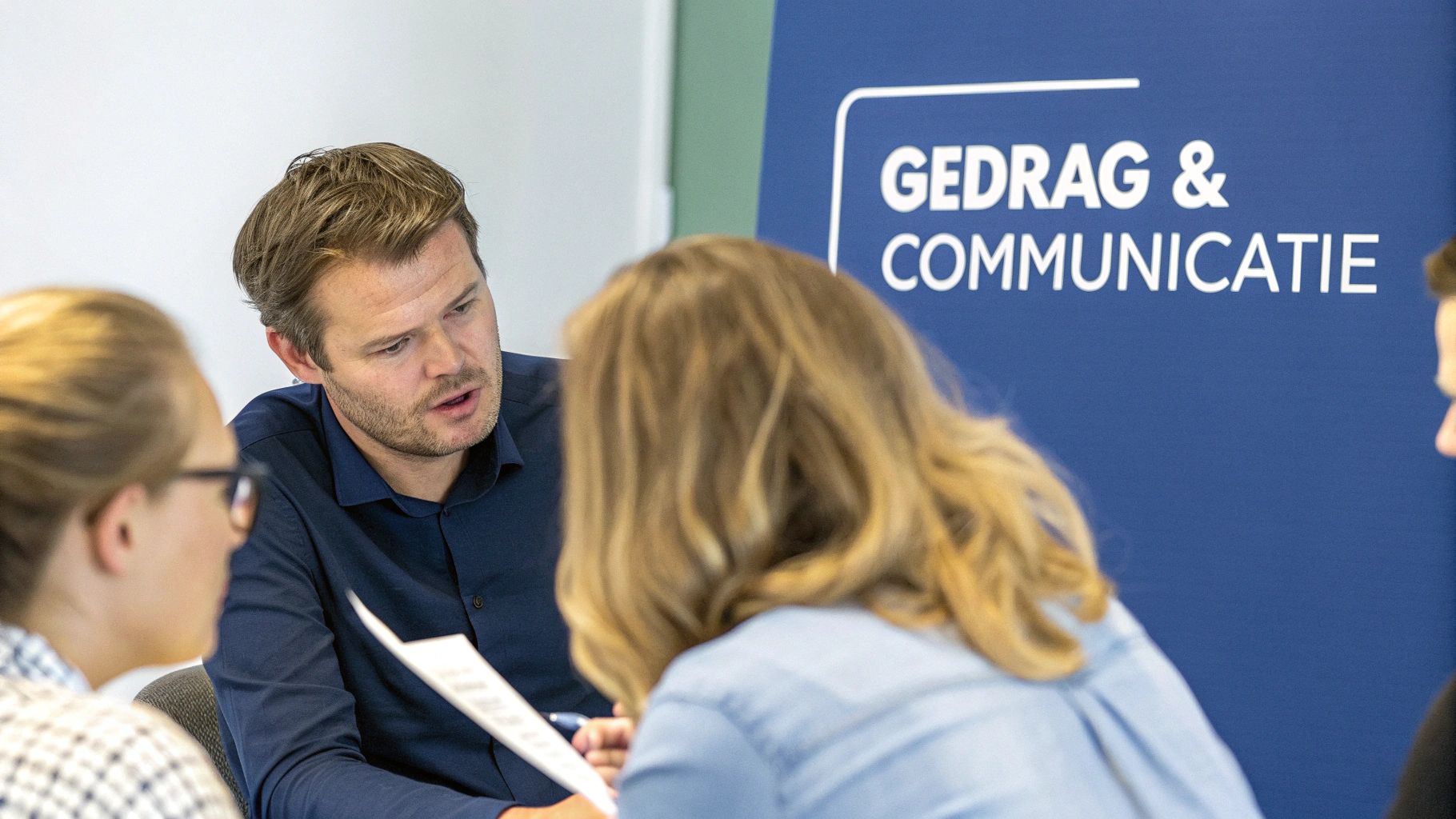 Man in blauw overhemd bespreekt documenten met twee vrouwen tijdens een training over gedrag en communicatie.