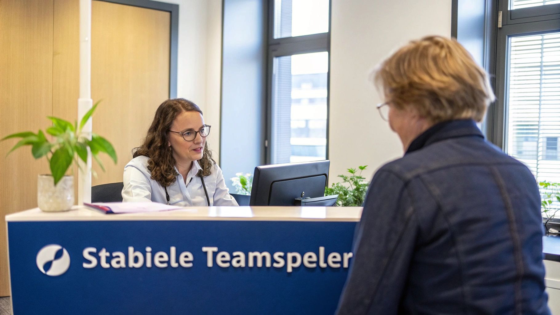 Vriendelijke receptioniste met bril spreekt een bezoeker aan een receptiebalie in een moderne kantoorruimte.