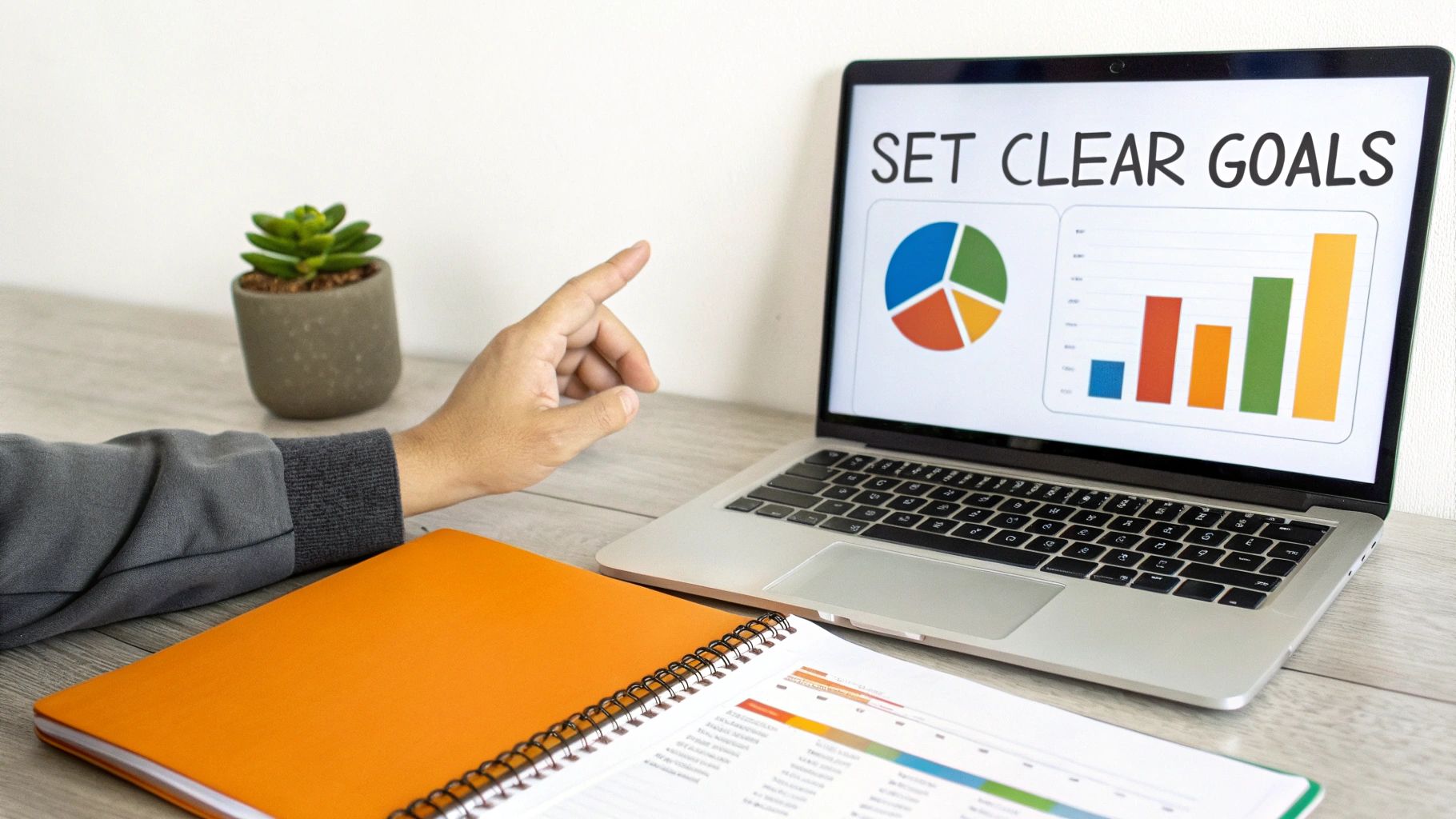 Hand pointing at laptop screen displaying 'SET CLEAR GOALS' with charts, next to a notebook.