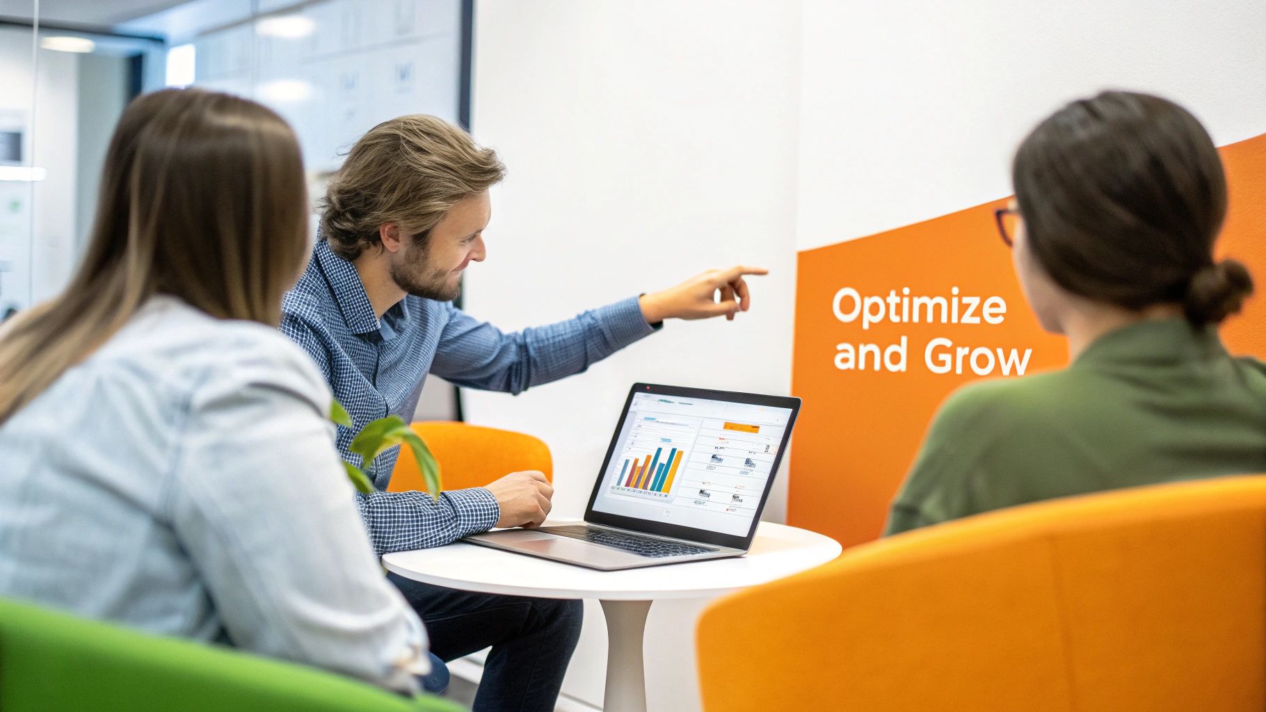 Smiling man points at laptop screen with business data to two colleagues in a vibrant office.