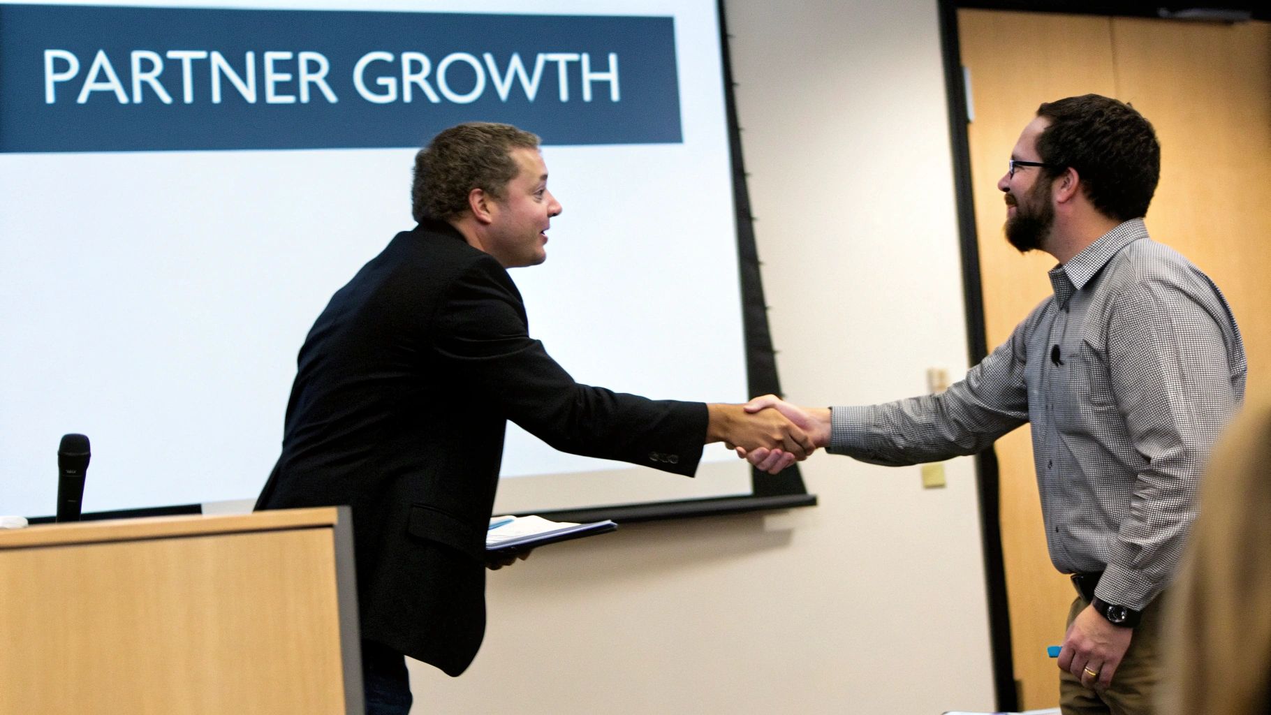 Two professional men shake hands on stage during a business presentation about partner growth.