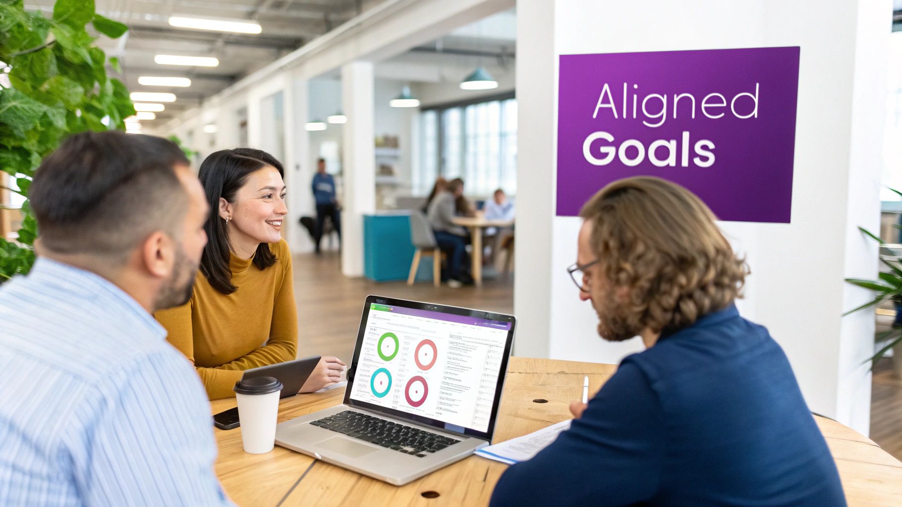 Three diverse professionals collaborate in a modern office, viewing data on a laptop, with an "Aligned Goals" sign.