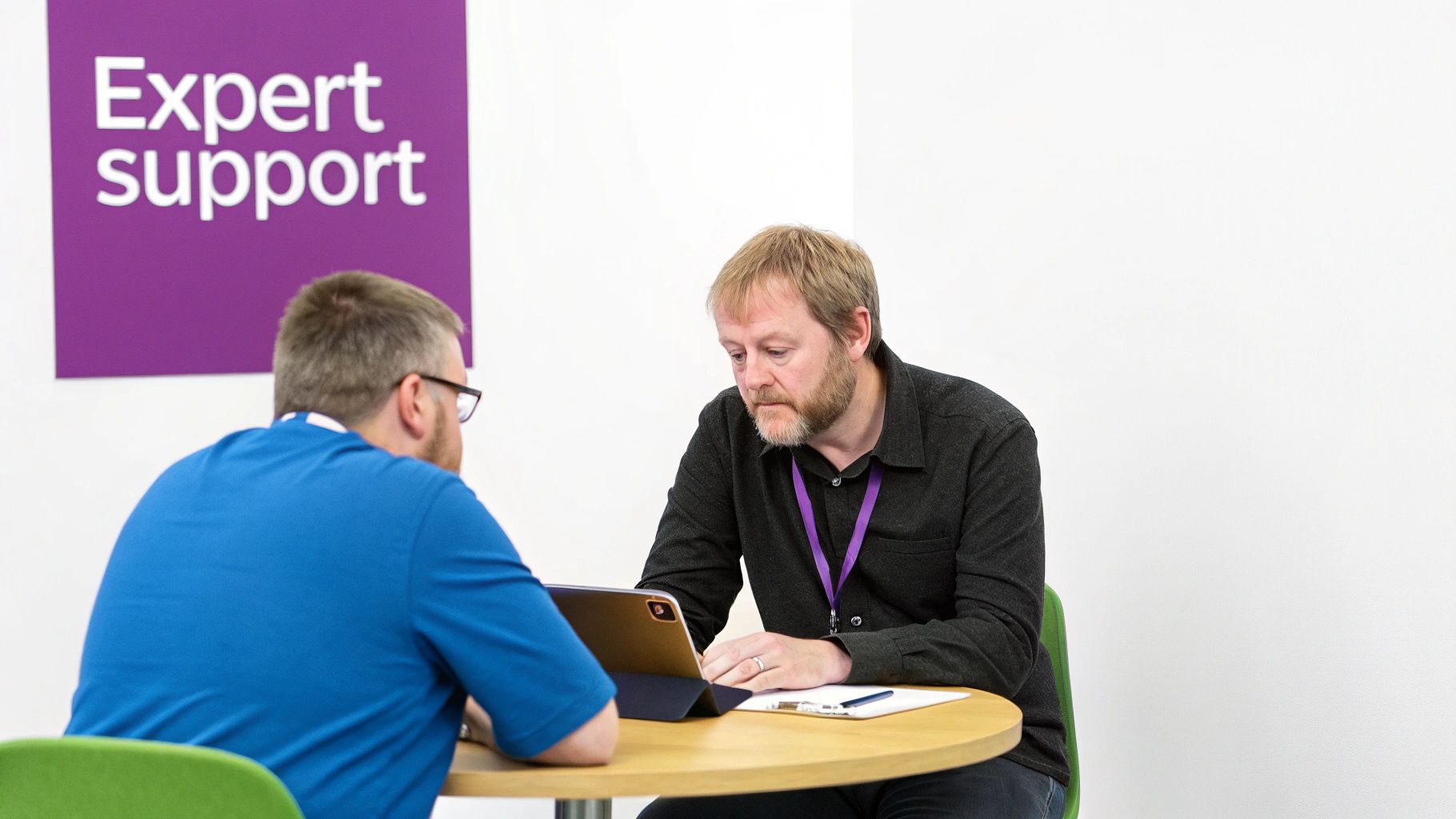 Two men sit at a table; one looks at a tablet, with an 'Expert support' sign visible.