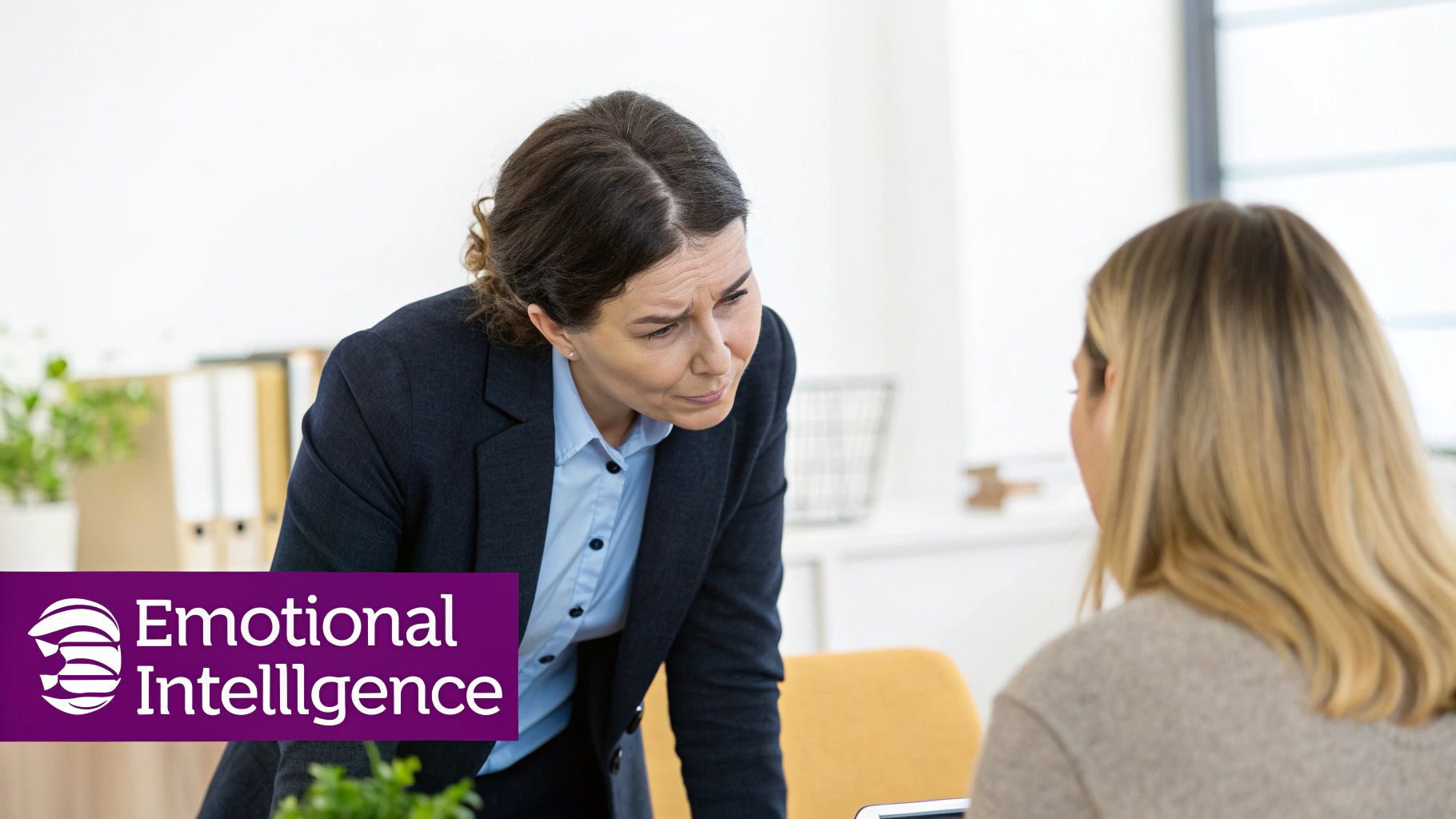 A serious-looking woman in a suit leans towards a blonde colleague in an office, with 'Emotional Intelligence' logo.
