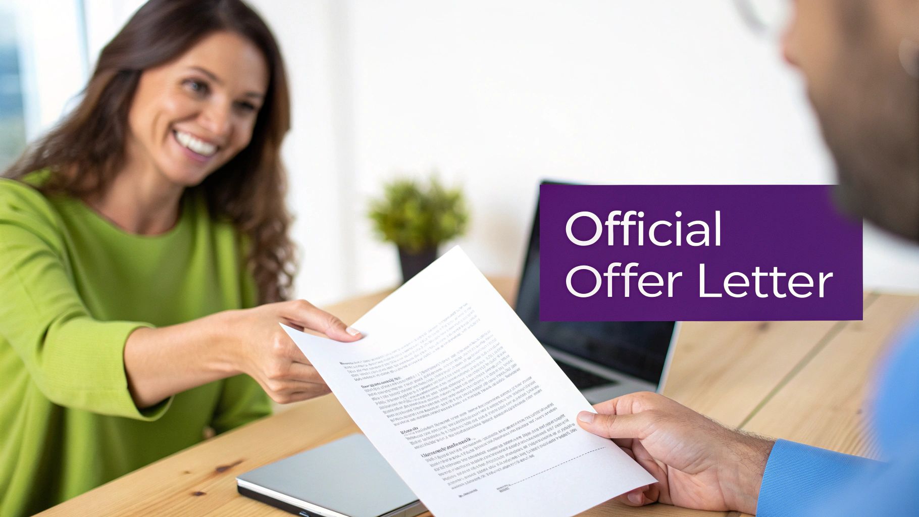 A smiling woman hands an official offer letter to a man across a wooden table.