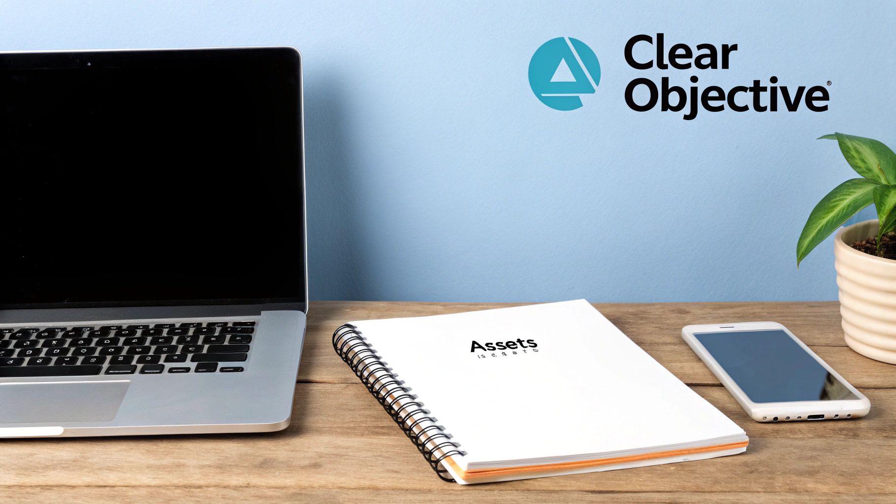 A modern office desk with a laptop, "Assets" notebook, smartphone, and a plant, with a "Clear Objective" logo on the blue wall.