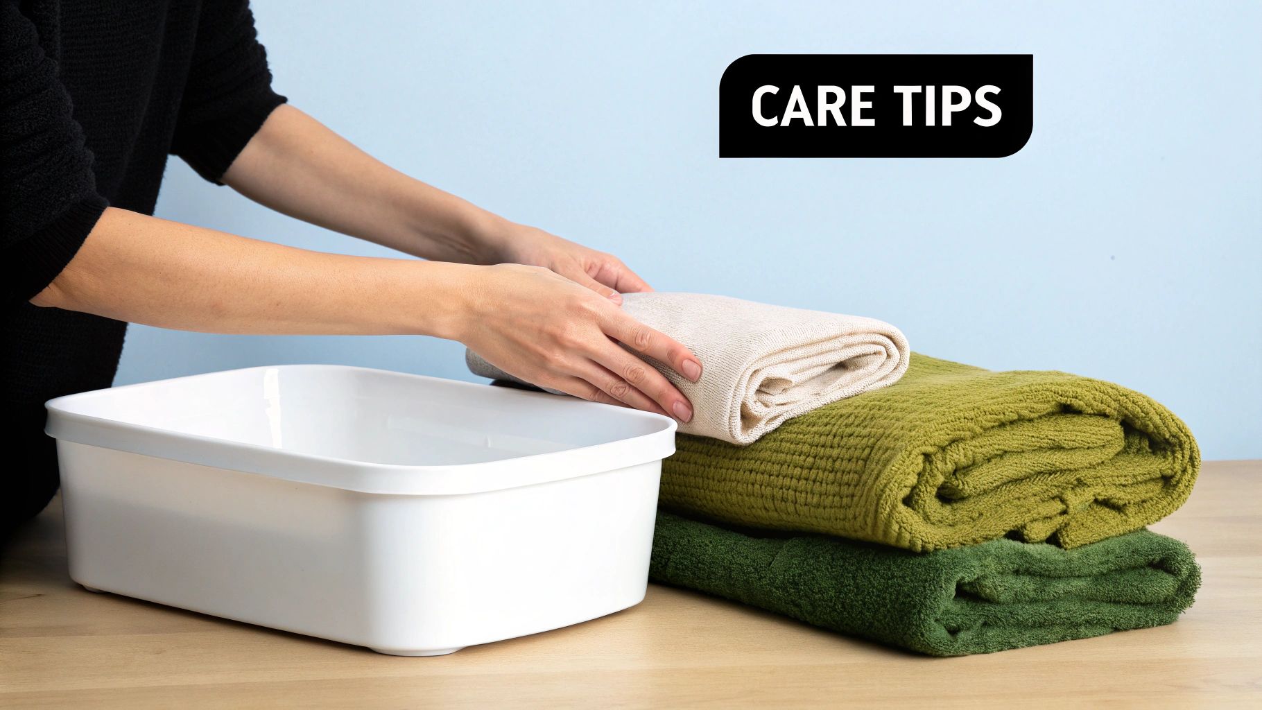 Person's hands folding a light-colored throw over a stack of green throws with a wash basin, illustrating care tips.