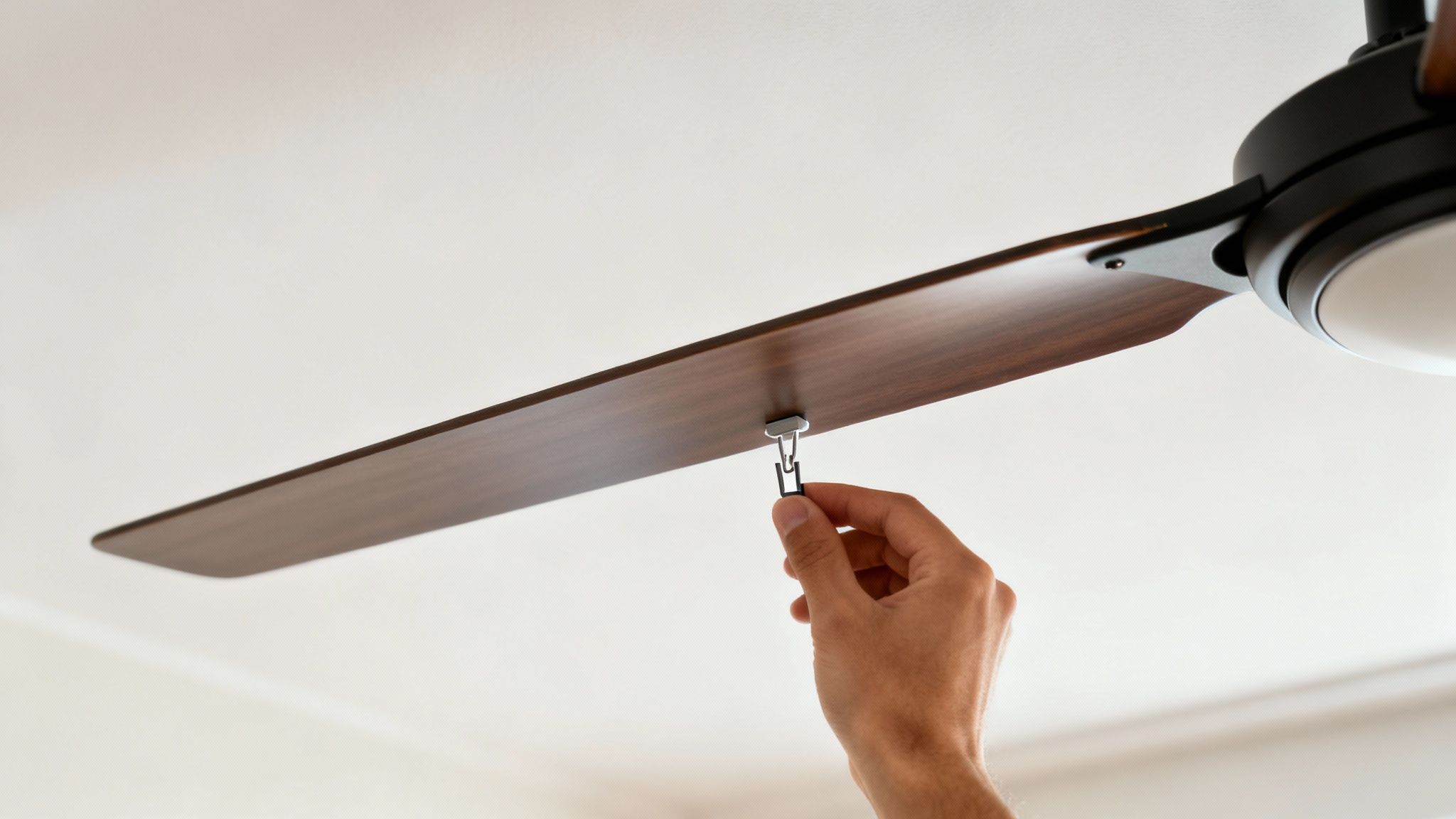 A hand adjusting a small silver and black balance clip on a dark wood ceiling fan blade.
