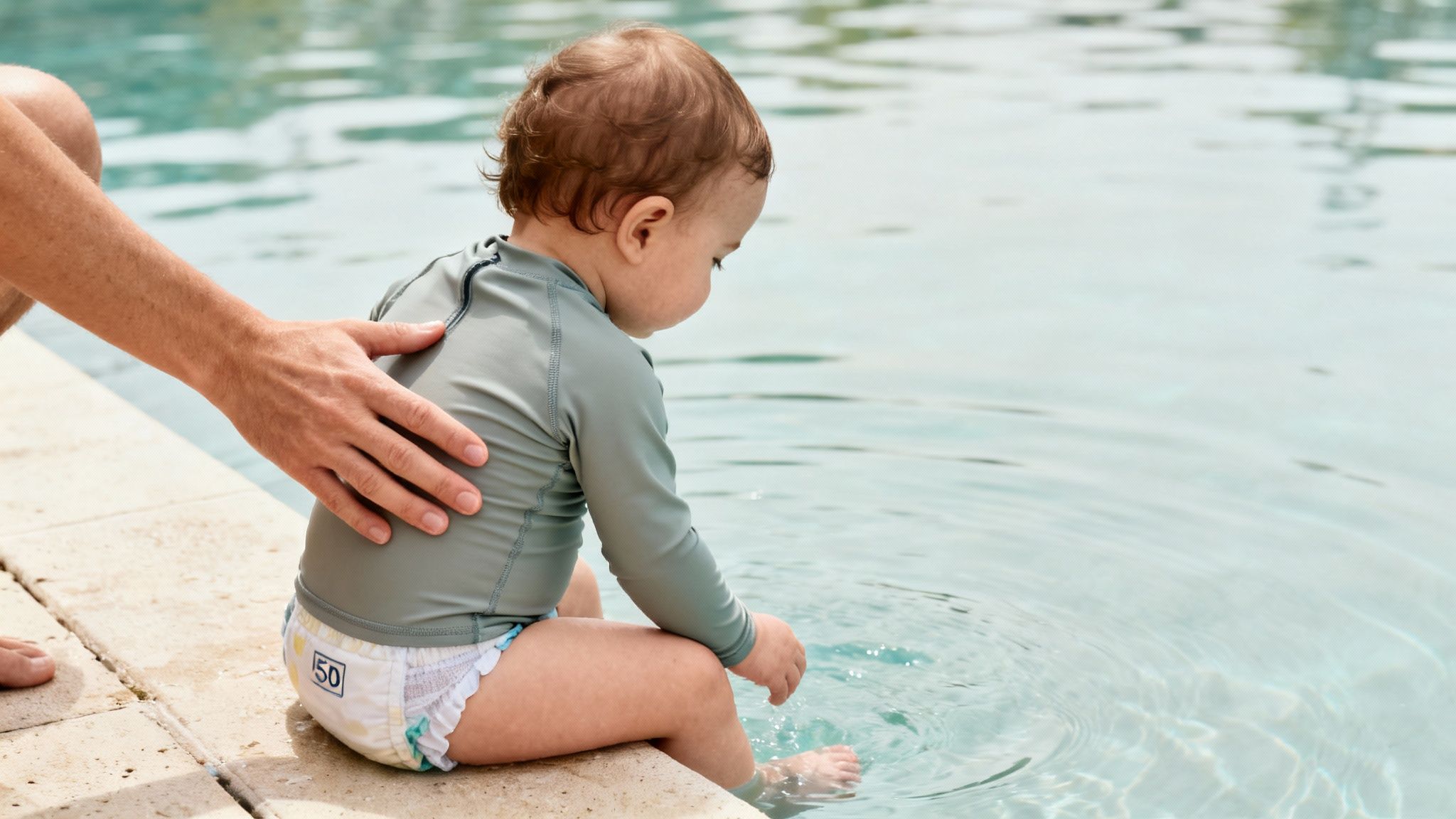 A baby boy, wearing a grey swim shirt and swim diaper, dips his feet into a swimming pool.