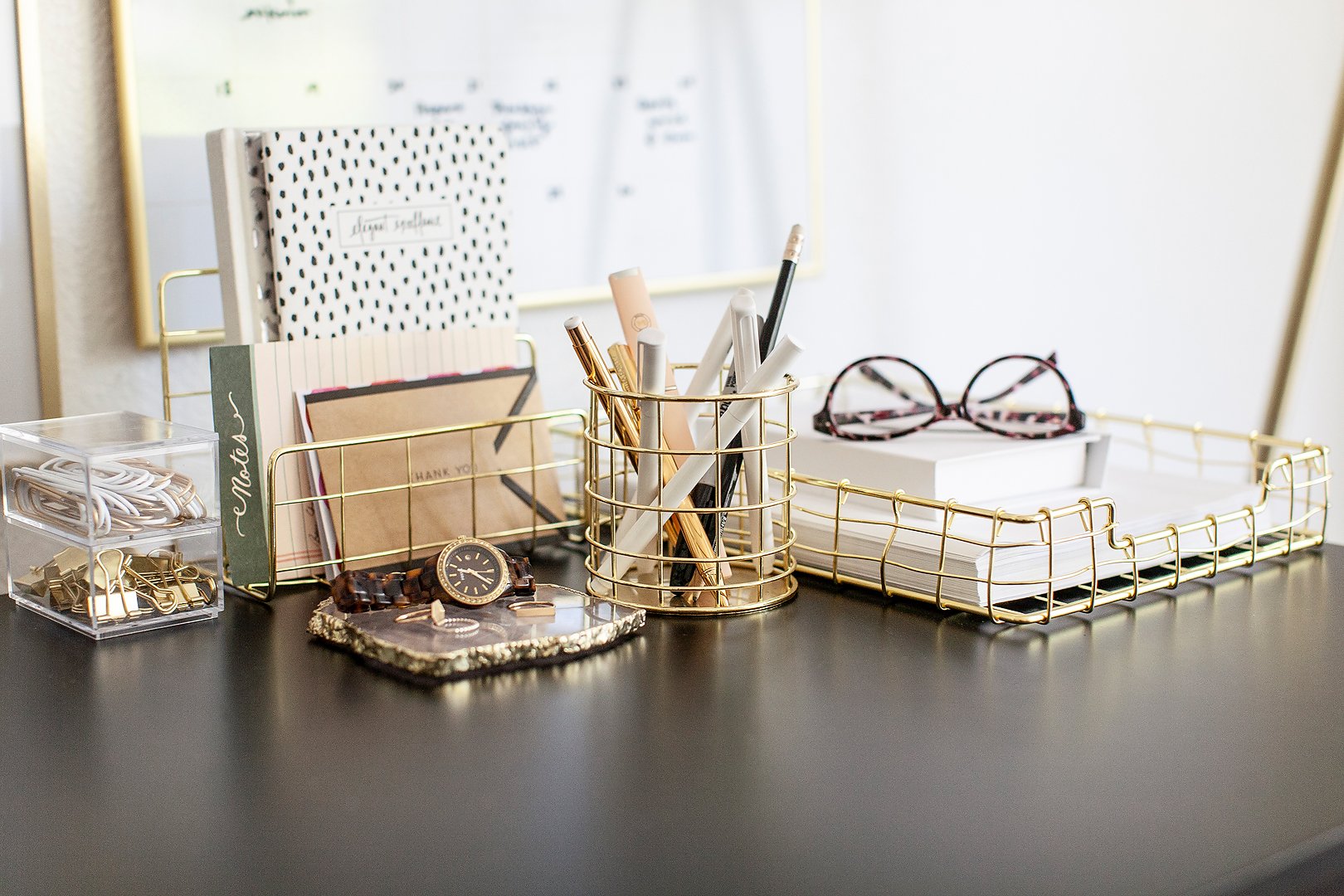 A stylishly organized desk with gold wire organizers, stationery, a watch, and glasses.