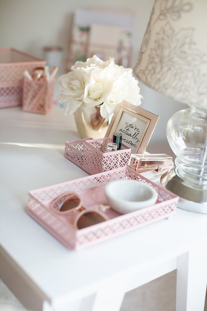 A neatly organized small desk featuring pink mesh trays, white flowers, and a motivational framed print.