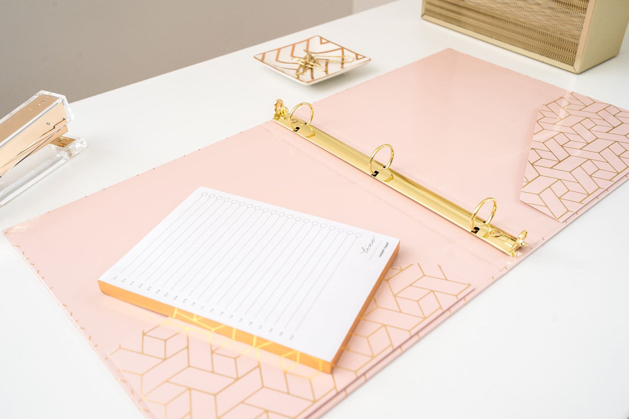 A stylish white desk with pink and gold office supplies, including a binder, stapler, and notepad.