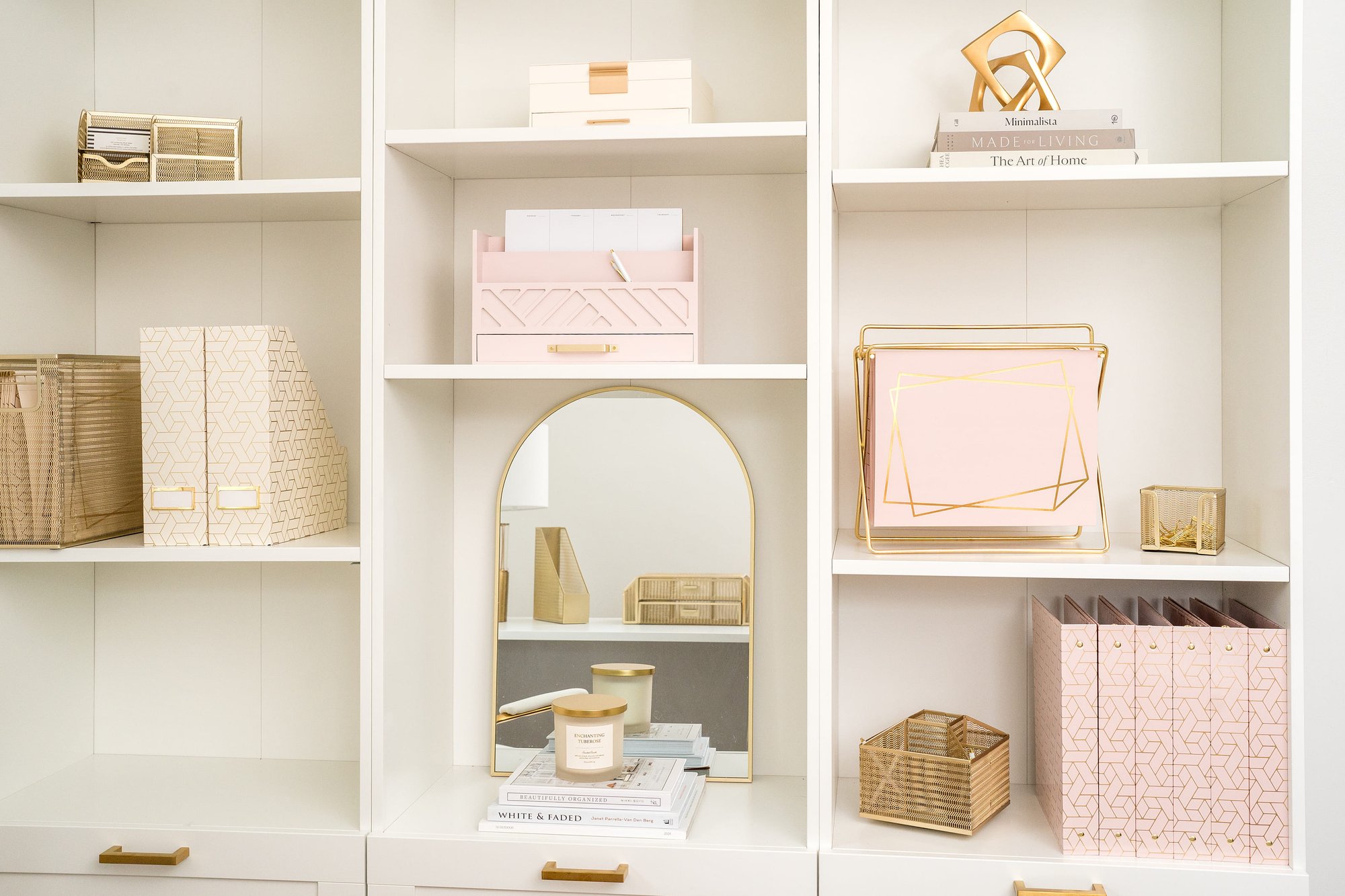 A beautifully organized white shelving unit displaying office supplies and decor in pink and gold tones.