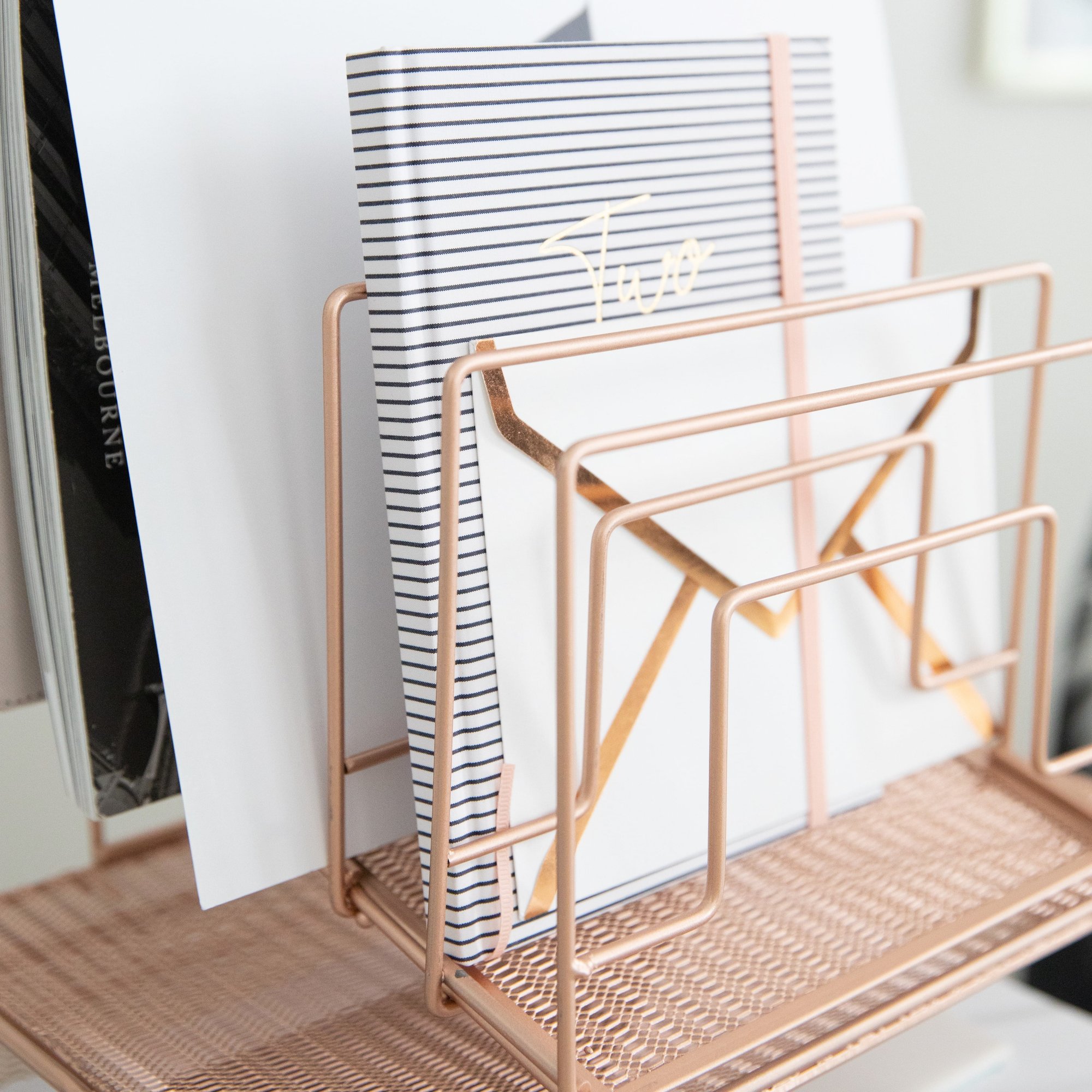A rose gold metal desk organizer holds a striped notebook and white envelopes, perfect for office decor.