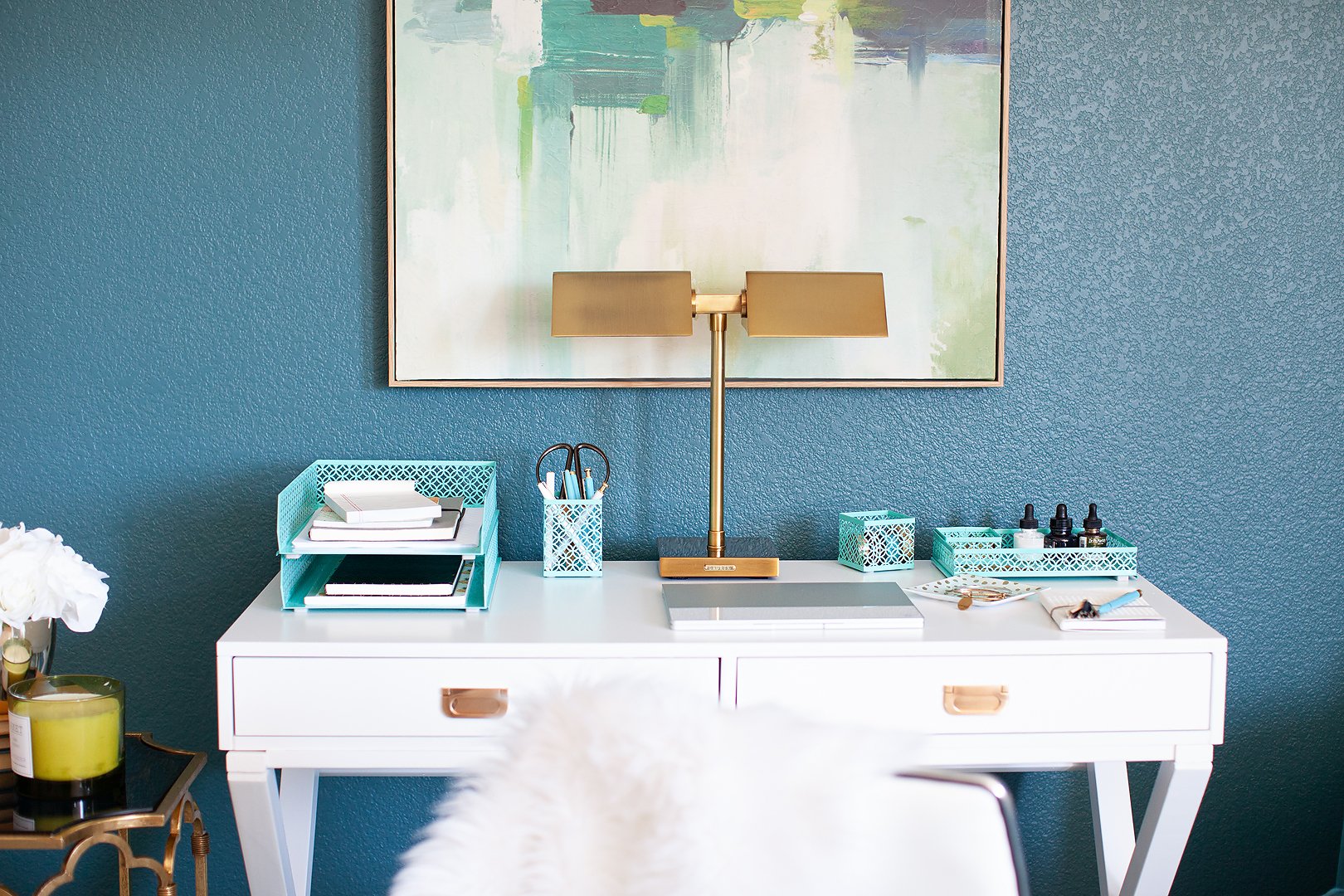 A stylish white desk with teal organizers, a gold lamp, and a laptop against a blue wall.