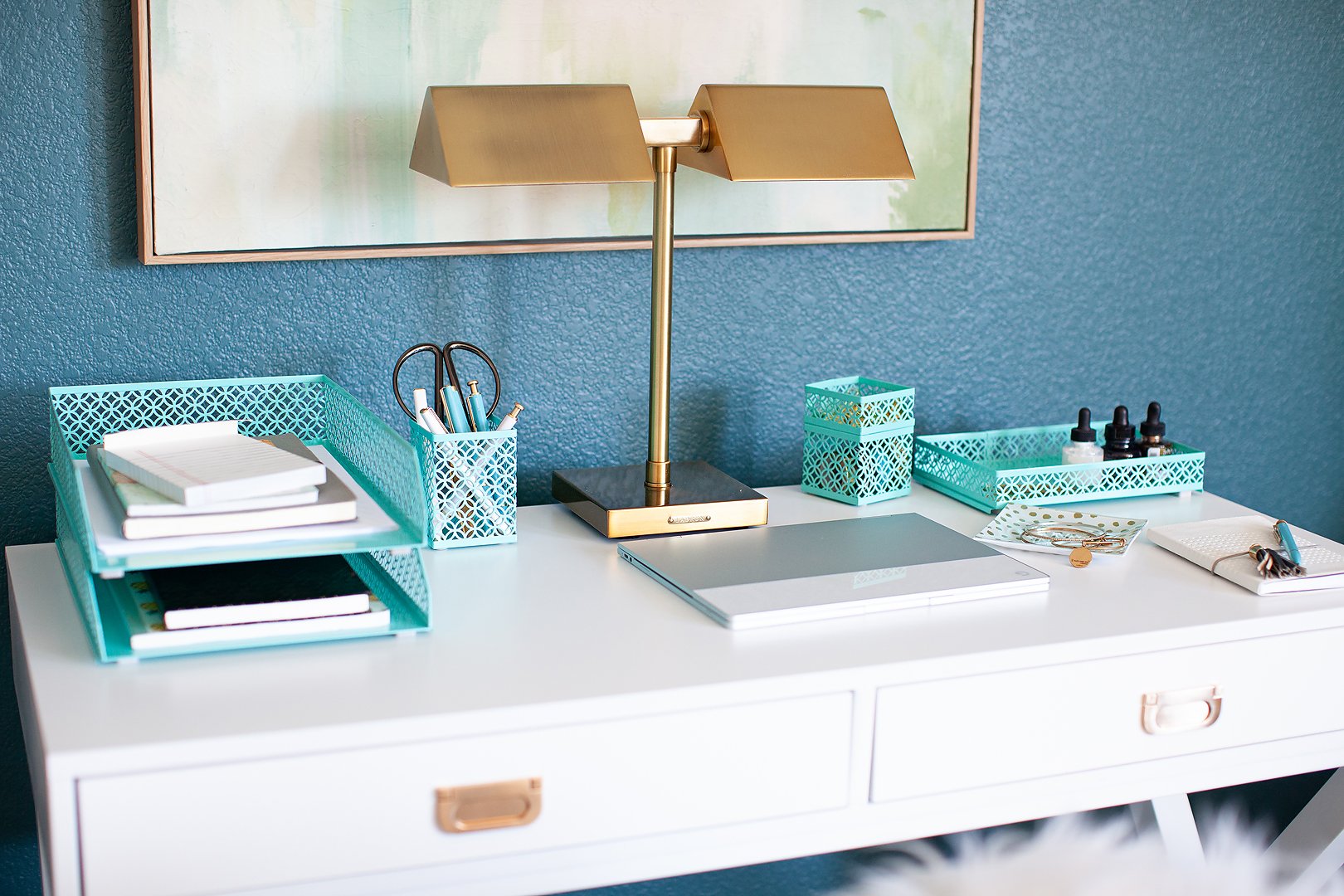 A neatly organized white desk featuring teal office supplies, a gold lamp, and a closed laptop.