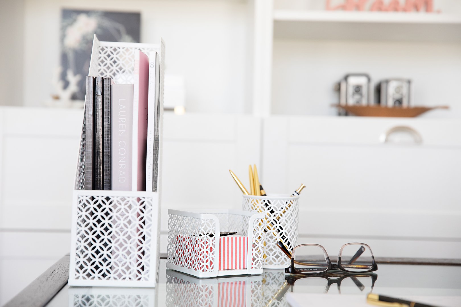 A clean white desk features decorative metal organizers holding books, gold pens, and small items, with reading glasses nearby.